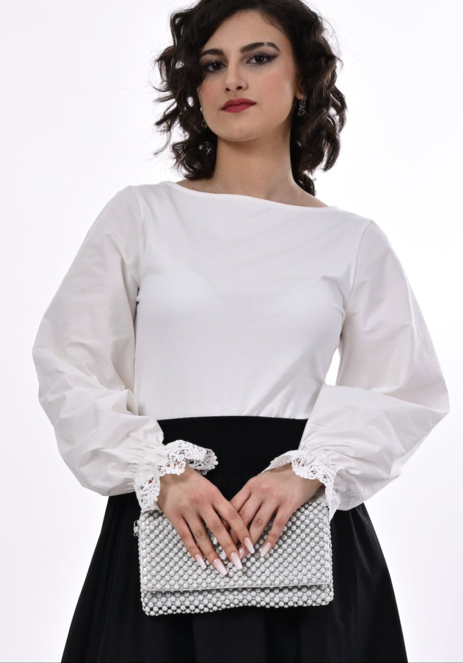 Woman wearing a white blouse with lace cuffs and a black skirt, holding a pearl clutch bag.