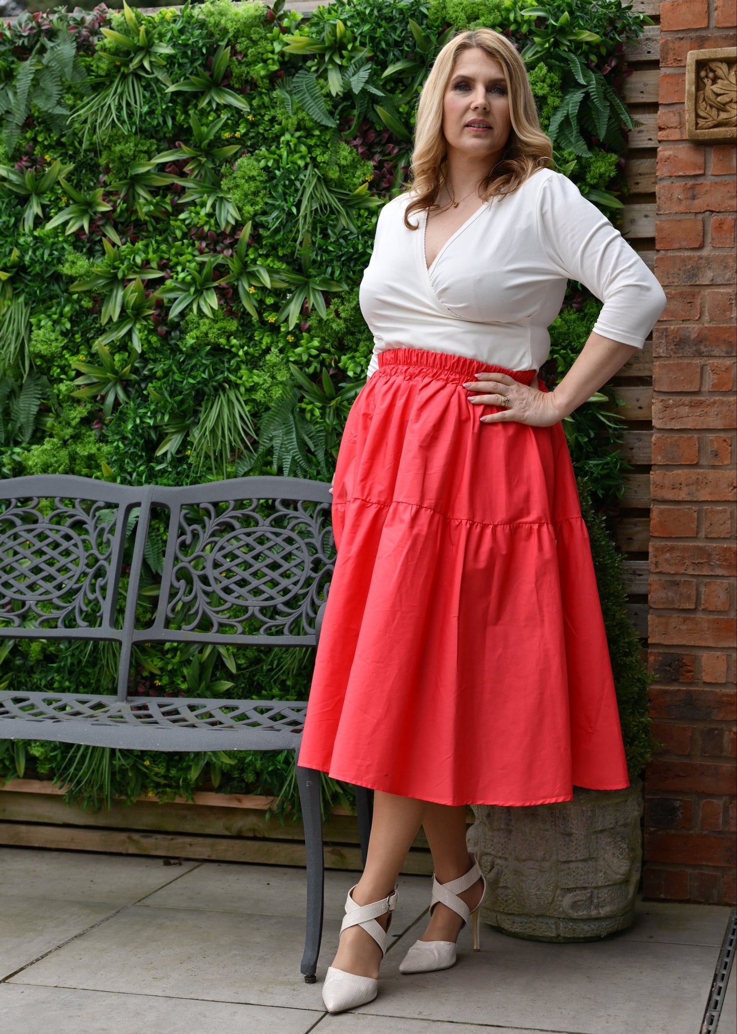A model wearing a midi length orangey red skirt with pockets, paired with a cream top and cream shoes, standing near a brick wall with greenery in the background.