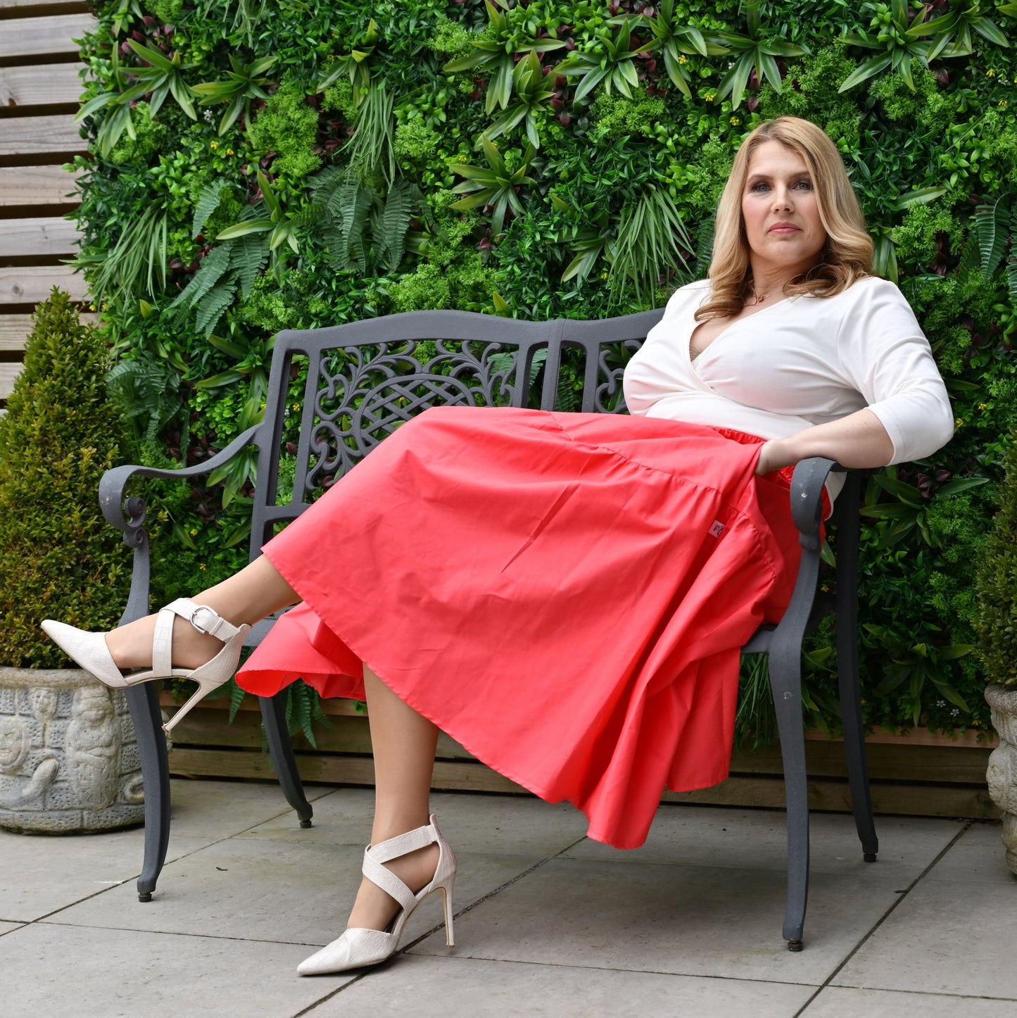 A model wearing a midi length orangey red skirt with pockets, paired with a cream top and cream shoes, sitting on a bench near a brick wall with greenery in the background.