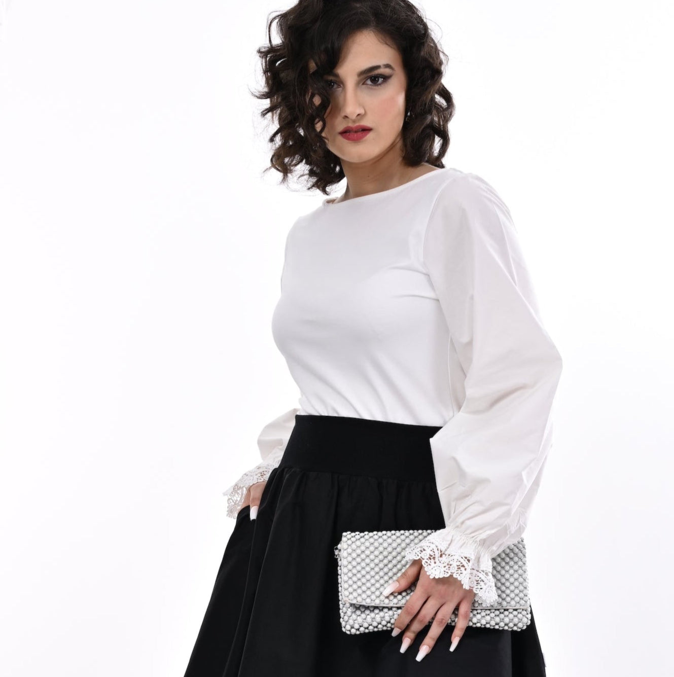 Woman wearing a white blouse with black skirt and pearl clutch bag on a white background.