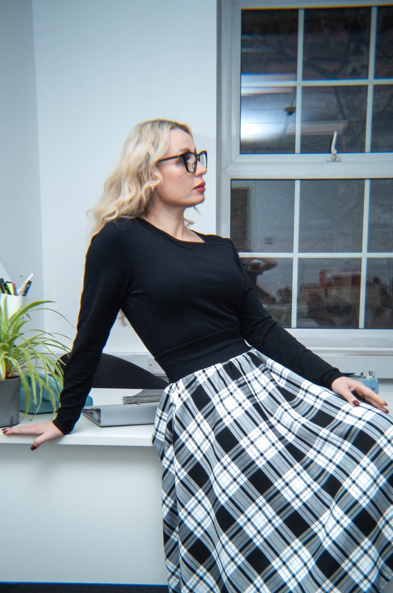 Woman sitting at a desk in an office setting wearing a black top and plaid skirt.