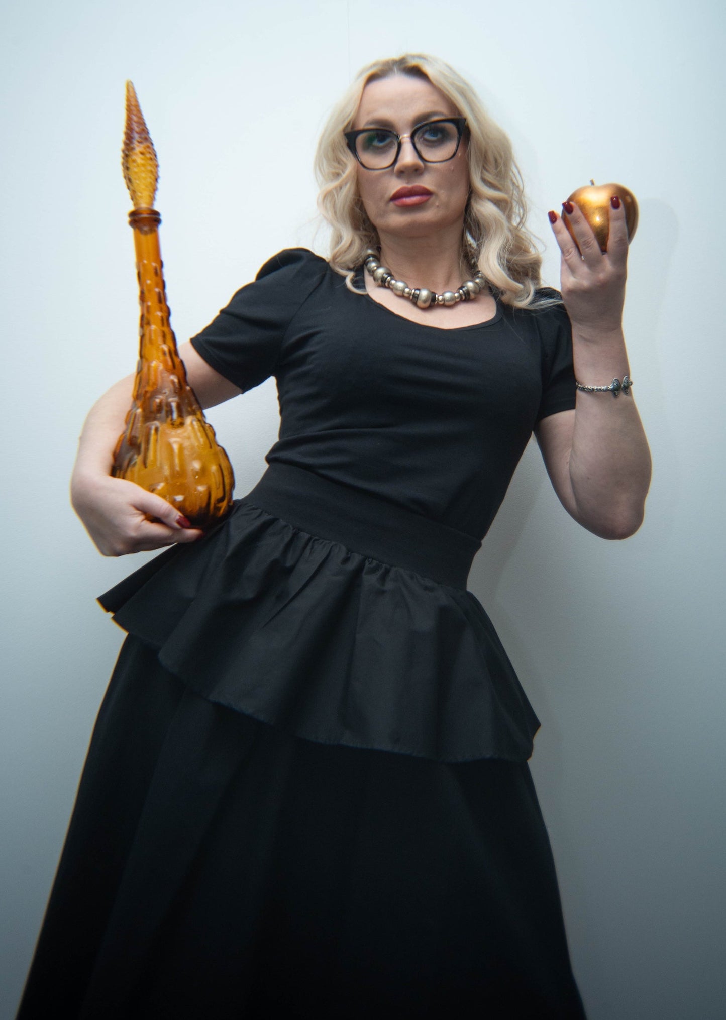 Woman in a black dress holding a large amber-colored pipe and an apple against a light blue background