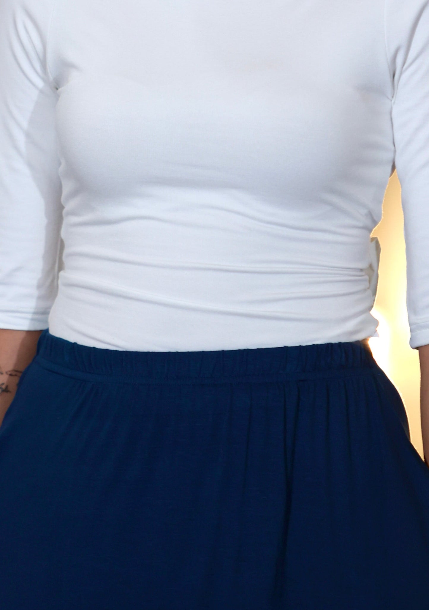 This is a young woman facing the camera silhouetted against a cream drape background. Her hair is tied back, she wears a cream 3/4 length sleeve top with a scoop neck. She wears a our blue Boho Skirt with is which has pockets and a deep gathered frill.
