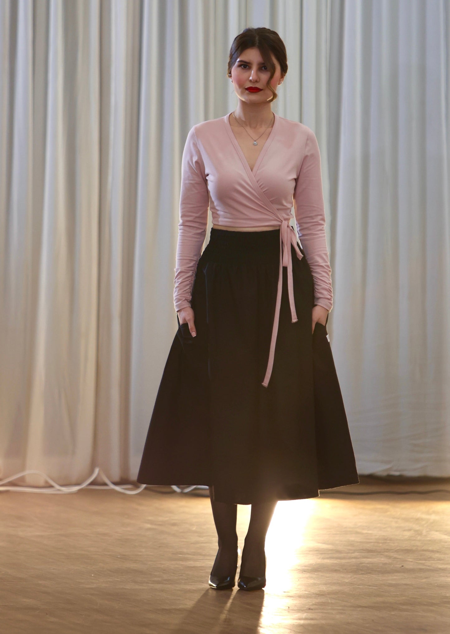 Woman wearing a pink top and black skirt against a white curtain background