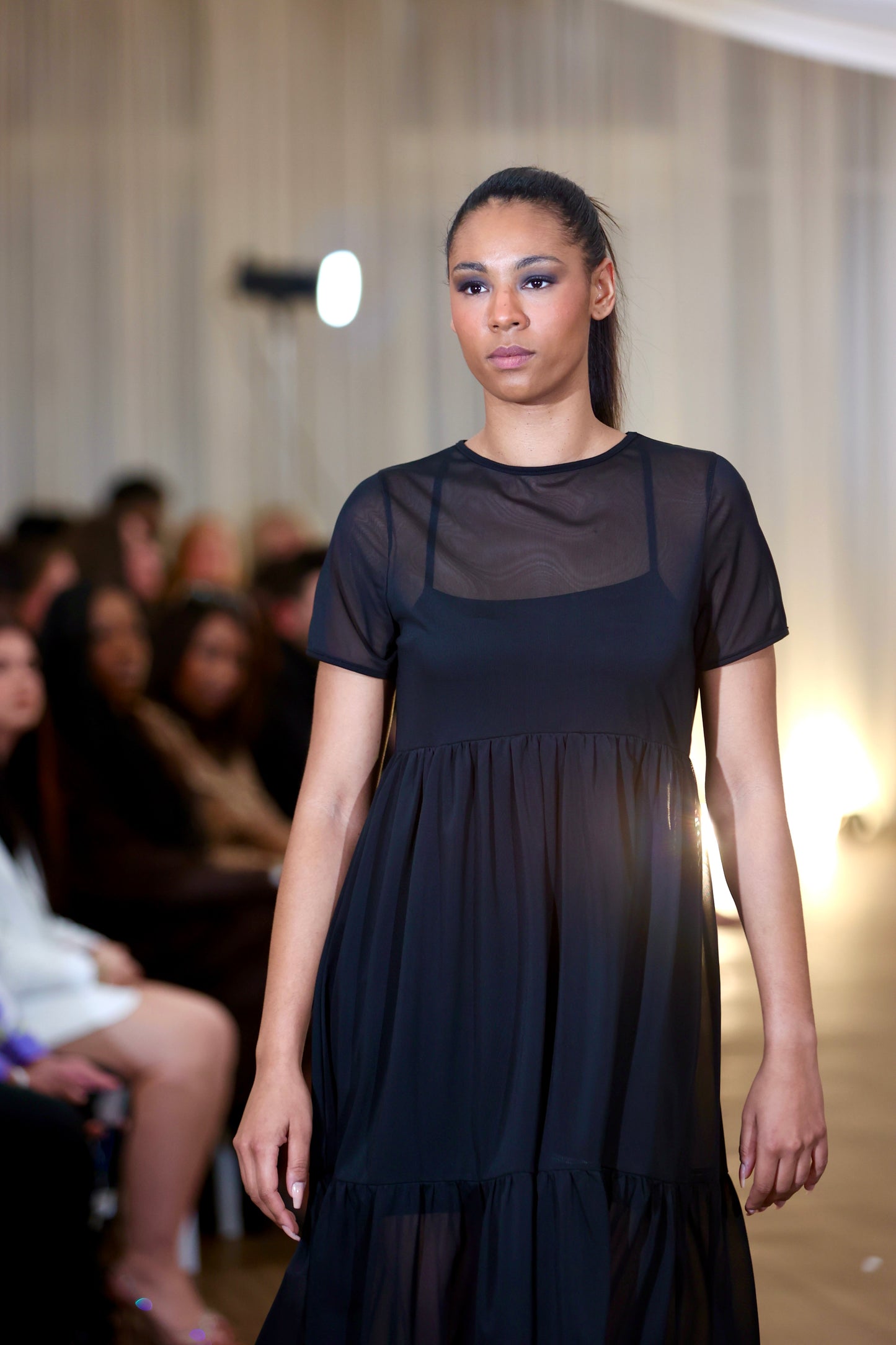 This is a young woman walking towards the cameral in a black mesh dress over our nectar cami dress. She is silhouetted against a blurred background of seated people. It has short sleeves and crew neck. There are waist gathers leading to a full skirt which has a deep gathered frill. This is a sheer dress, midi length.