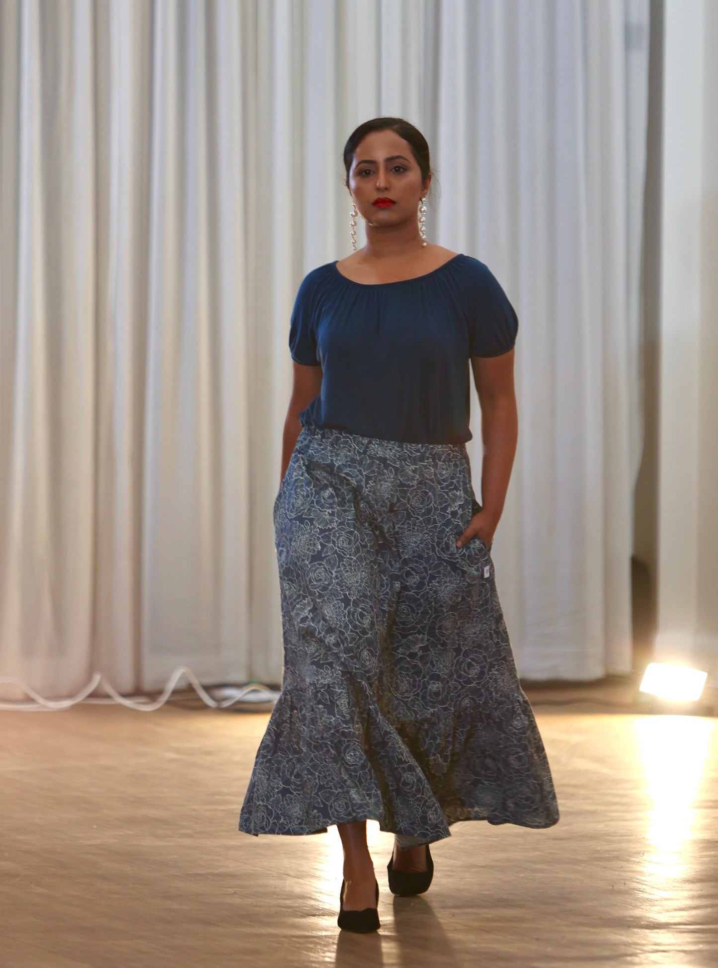 The young woman in the picture has dark hair and she is walking towards the camera in a well lit room. She is wearing a boho gathered midi skirt with pockets in navy print. She is also wearing our posey top in french navy. She is wearing black shoes. Her right hand is behind her back while her left hand is in a pocket.