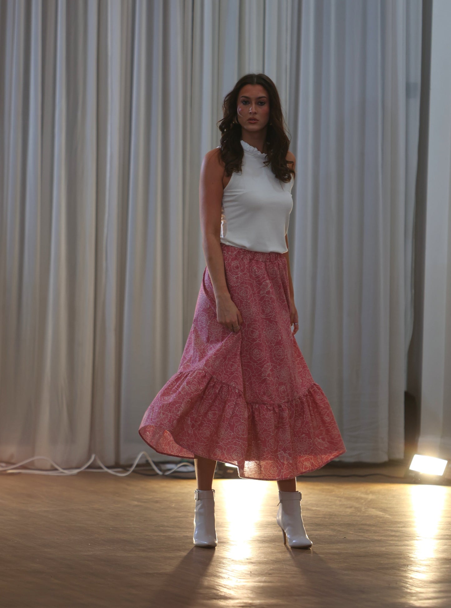 This is a young woman walking towards the camera., she has long dark hair. She is wearing a pink and cream patterned skirt silhouetted against a white drape background. It has an elasticated waist. She wears this with a cream sleeveless top with ruffle neck and she wears cream boots.