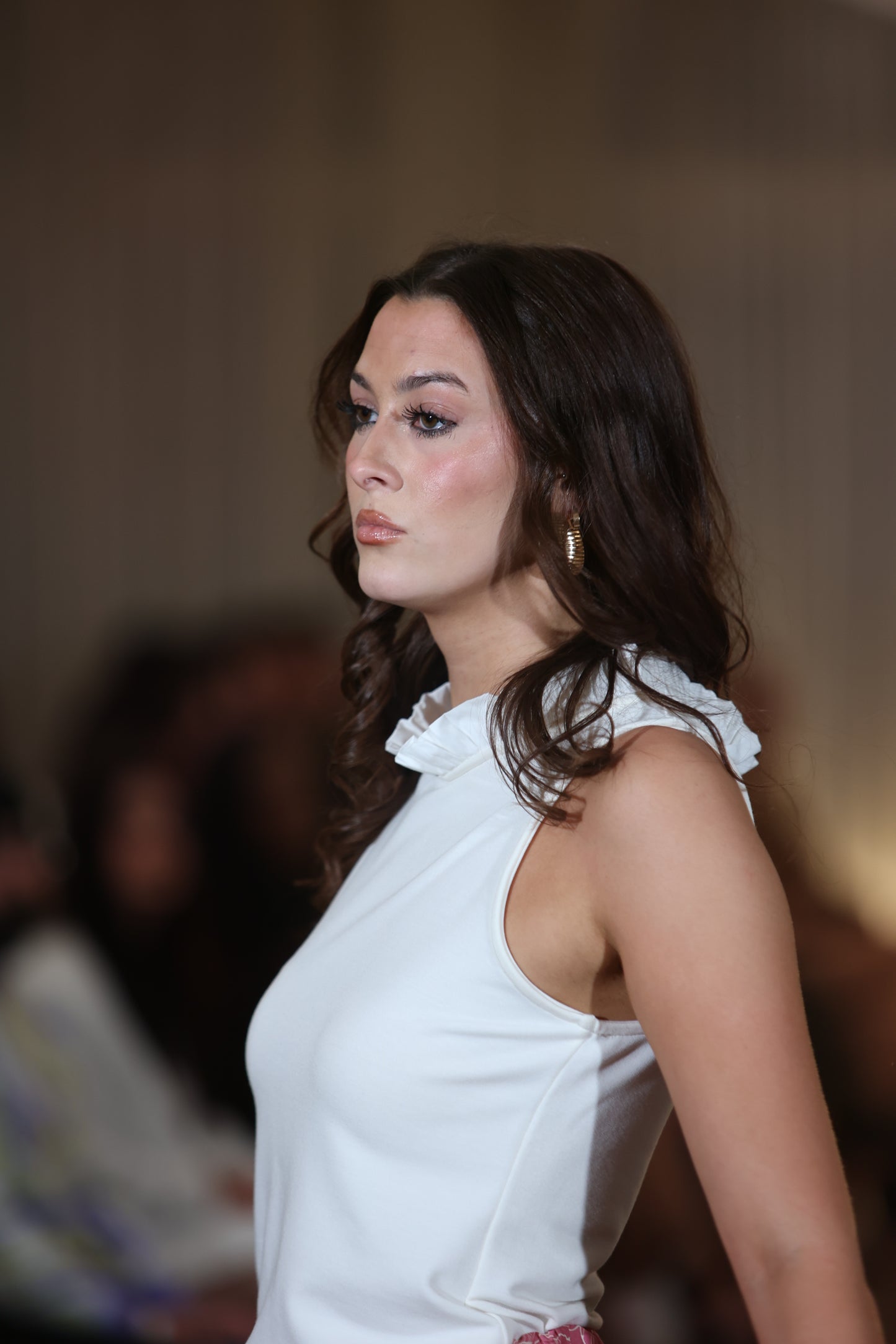 This is a young woman standing facing the left of the picture., she has long dark hair. She is wearing cream sleeveless top and is silhouetted against a neutral blurred background.
