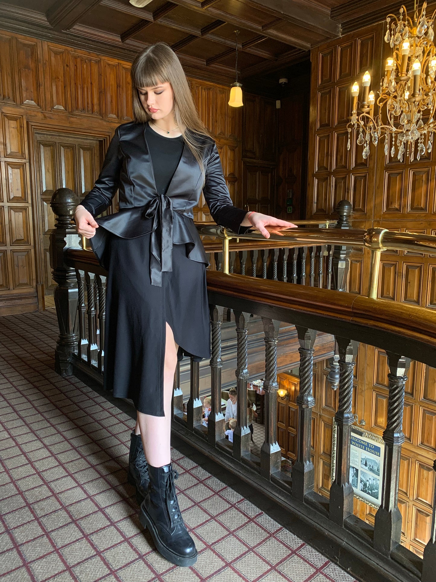 Our model faces the front but is looking down at the hem of our masquerade black satin jacket. She is holding the banister with her left hand and the hem of the jacket in her right hand. Her long dark hair is falling behind her down her back. She is on a landing with dark wood panelling and a brass banister. She is wearing a mock wrap skirt that has an asymmetrical hemline with a black crew neck long sleeved bodysuit.