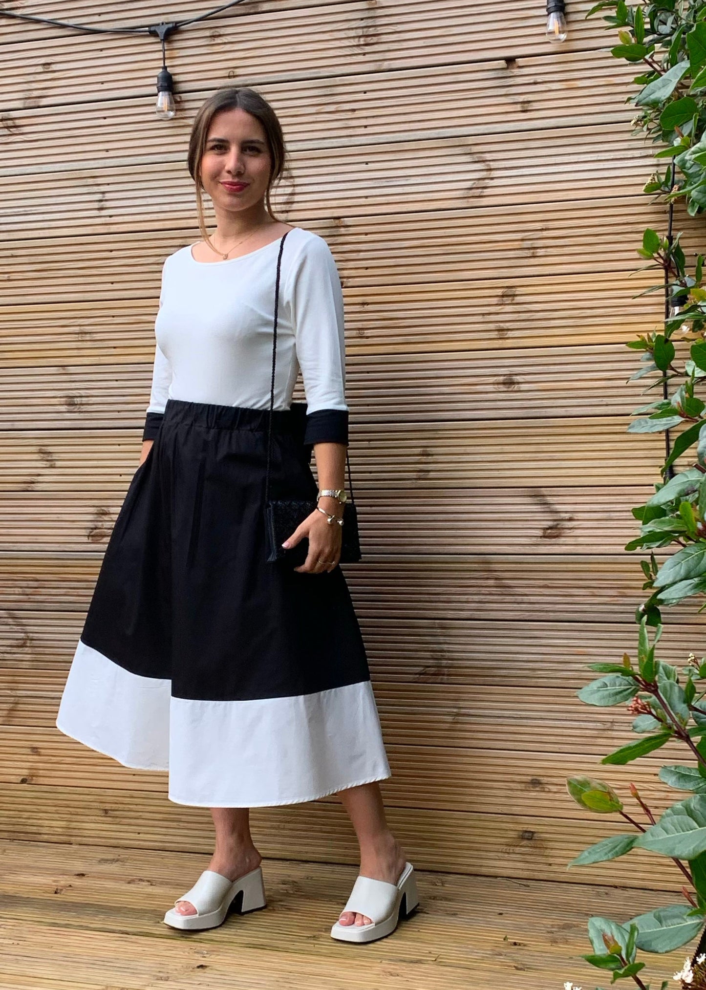A woman wearing a color-block midi skirt with a white top and white heeled sandals, standing in front of a wooden slat wall.