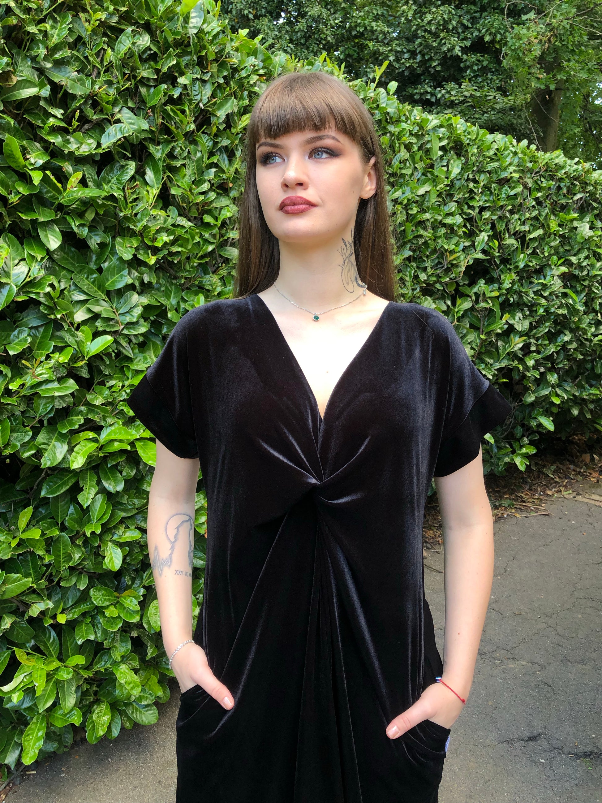 This is a black frill velvet dress worn by a model with dark hair against a Japonica hedge. The image is cut off at the top of her legs. It has a knot and plunge v neck with short sleeves. The model is facing front and has her hands in her pockets.