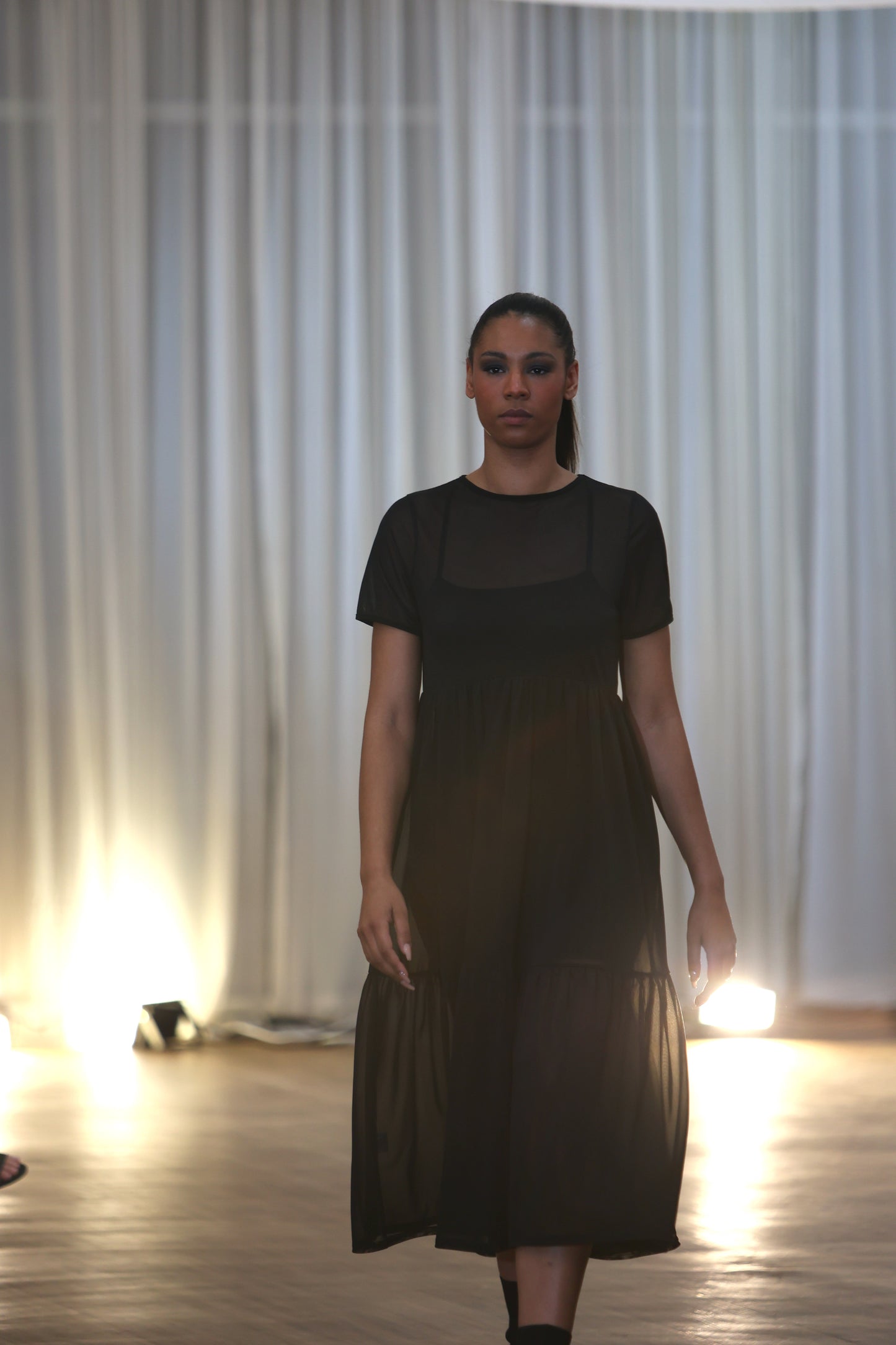This is a young woman walking towards the cameral in a black mesh dress over our nectar cami dress. She is silhouetted against a blurred background of seated people. It has short sleeves and crew neck. There are waist gathers leading to a full skirt which has a deep gathered frill. This is a sheer dress, midi length.