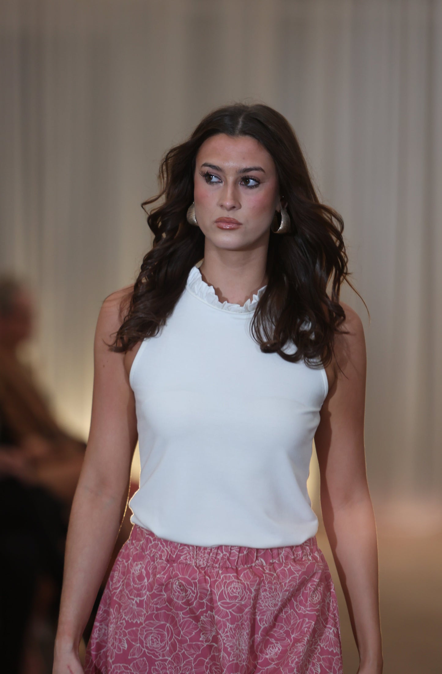 This is a young woman walking towards the camera., she has long dark hair. She is wearing a pink and cream patterned skirt silhouetted against a white drape background. It has an elasticated waist. She wears this with a cream sleeveless top with ruffle neck.