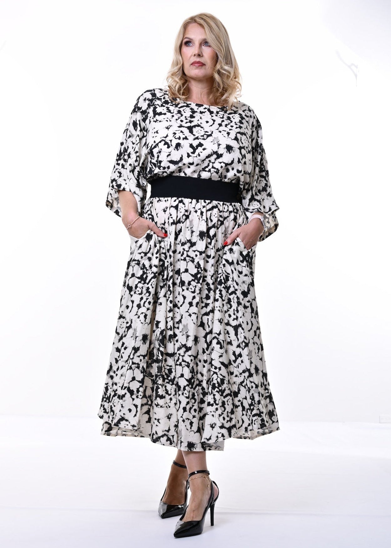 A woman modeling a black and white abstract patterned midi dress with a black waist belt, standing against a plain white studio background.