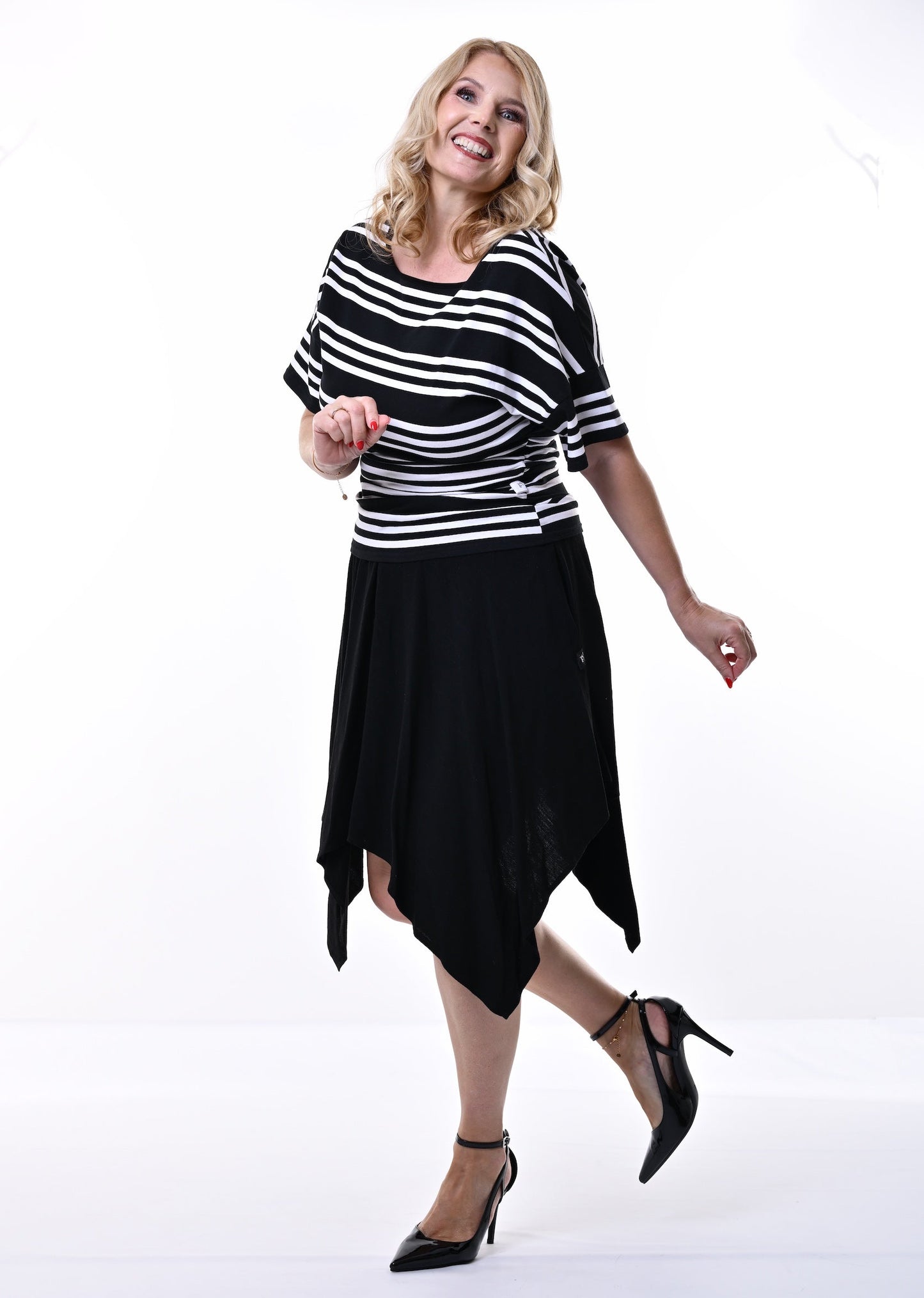 A woman modeling a black and white horizontally striped short-sleeve top with a black high-low skirt and black heels, posing in a bright white studio setting.