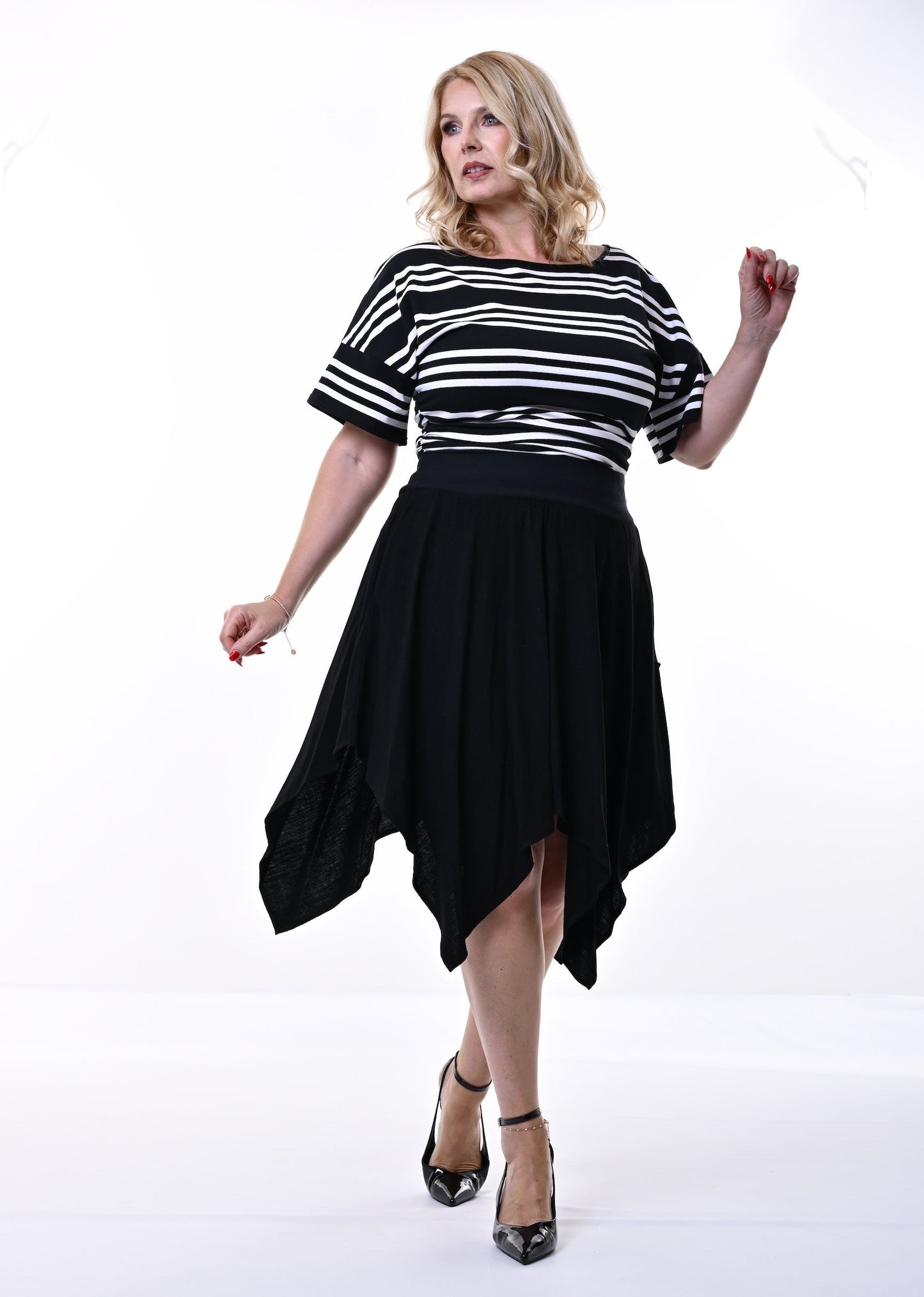 A woman modeling a black and white horizontally striped short-sleeve top with a black high-low skirt, paired with black pointed-toe heels, against a white studio background.