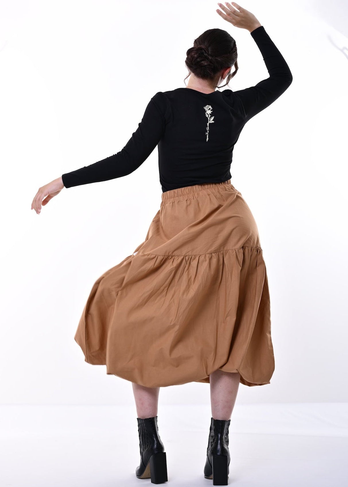 A woman is shown from behind wearing a black long-sleeve top with a small white graphic on the back and a tiered brown skirt, posing in a bright white studio setting.
