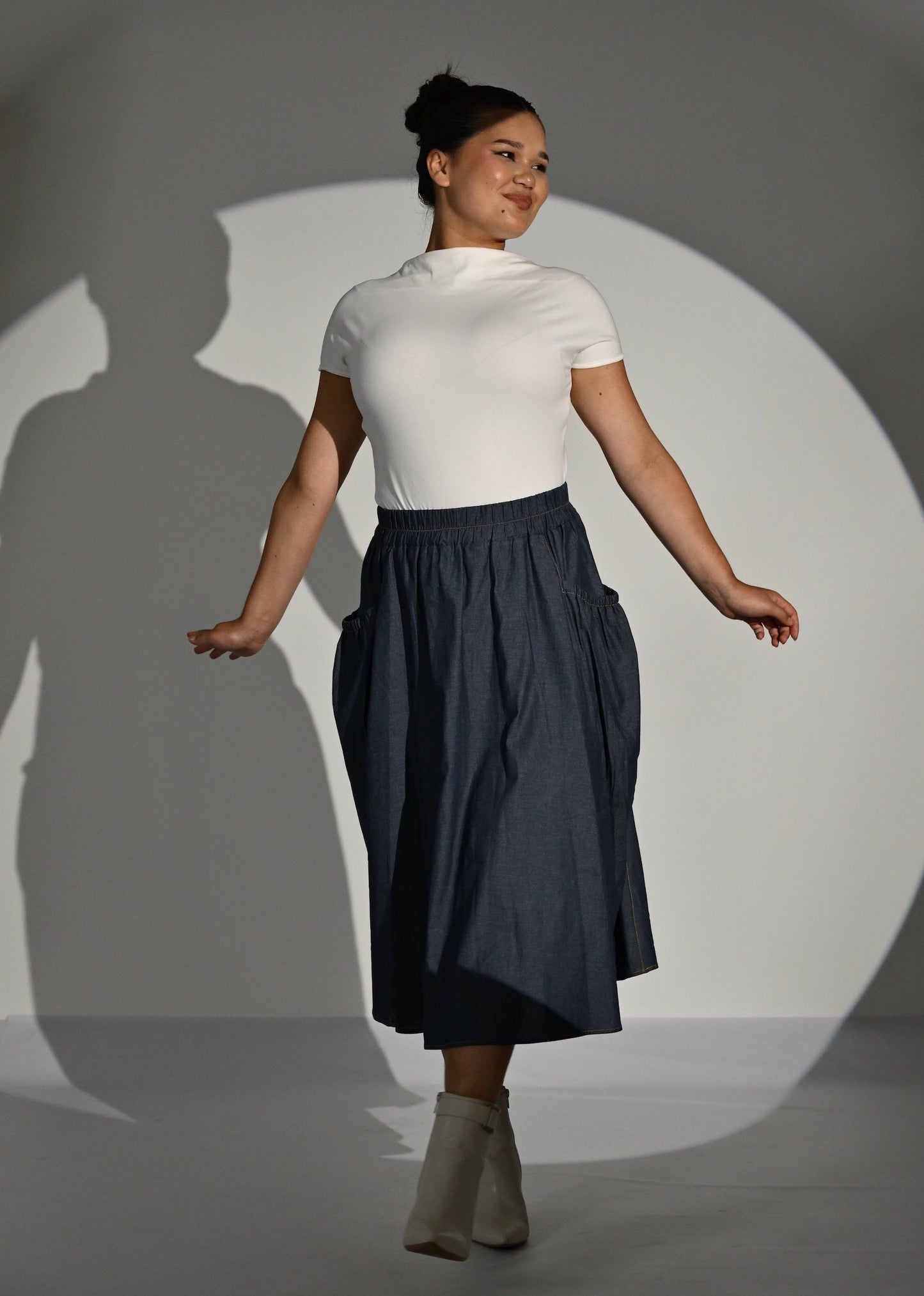 A woman modelling a cream short-sleeve mock neck top and a dark navy blue high-waisted pleated midi skirt, paired with beige ankle boots, set against a gray studio backdrop with a spotlight and shadow.