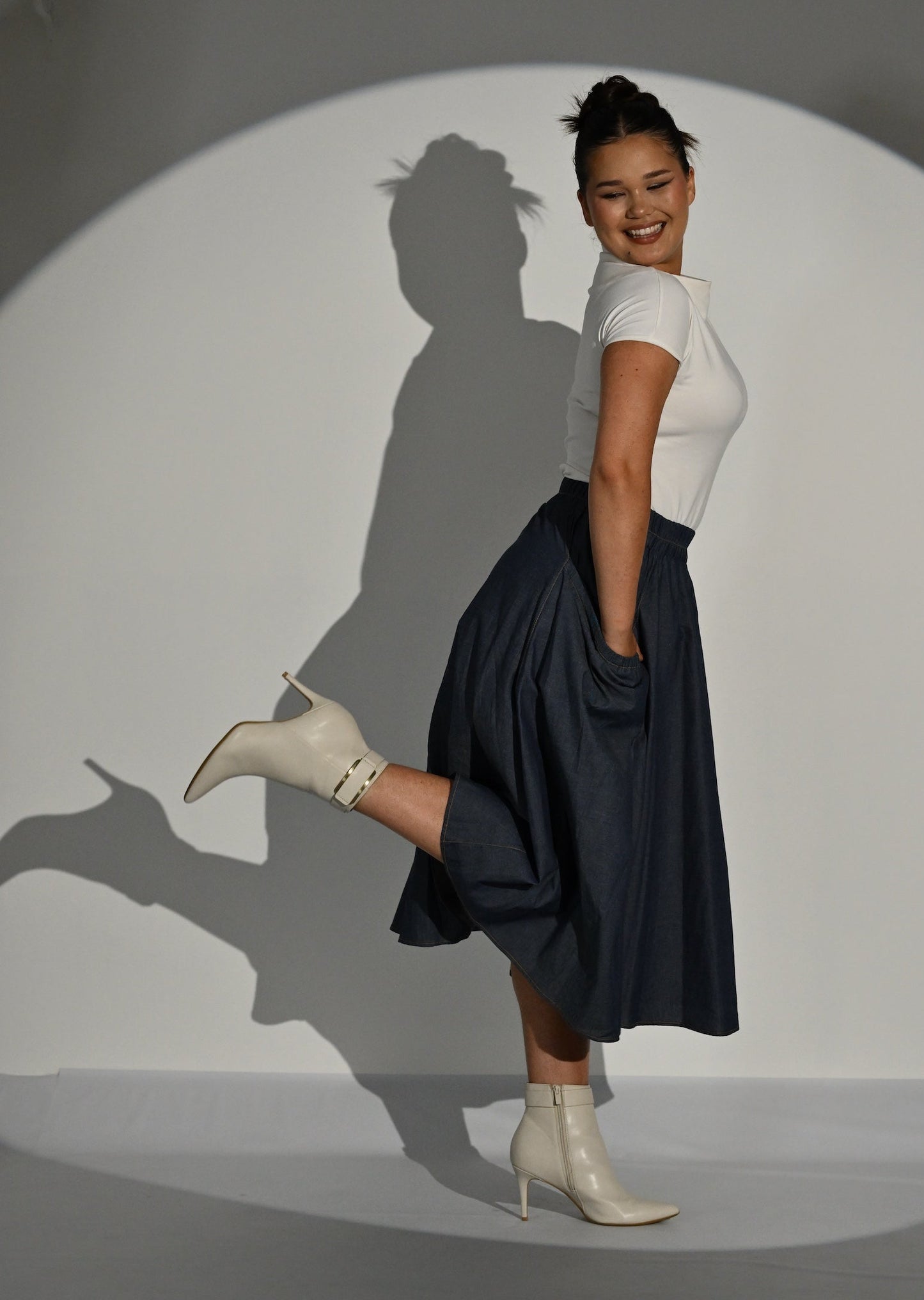 A woman posing in a studio setting under a spotlight, wearing a cream short-sleeve top, a dark navy midi skirt, and white ankle boots.