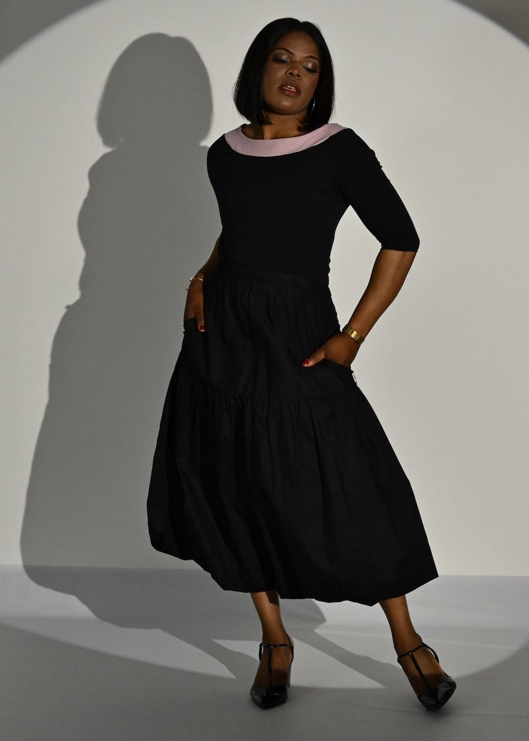 A woman modelling a black top with a light pink collar, standing in front of a white spotlight on a gray studio floor. The spotlight casts a long shadow on the wall behind her.