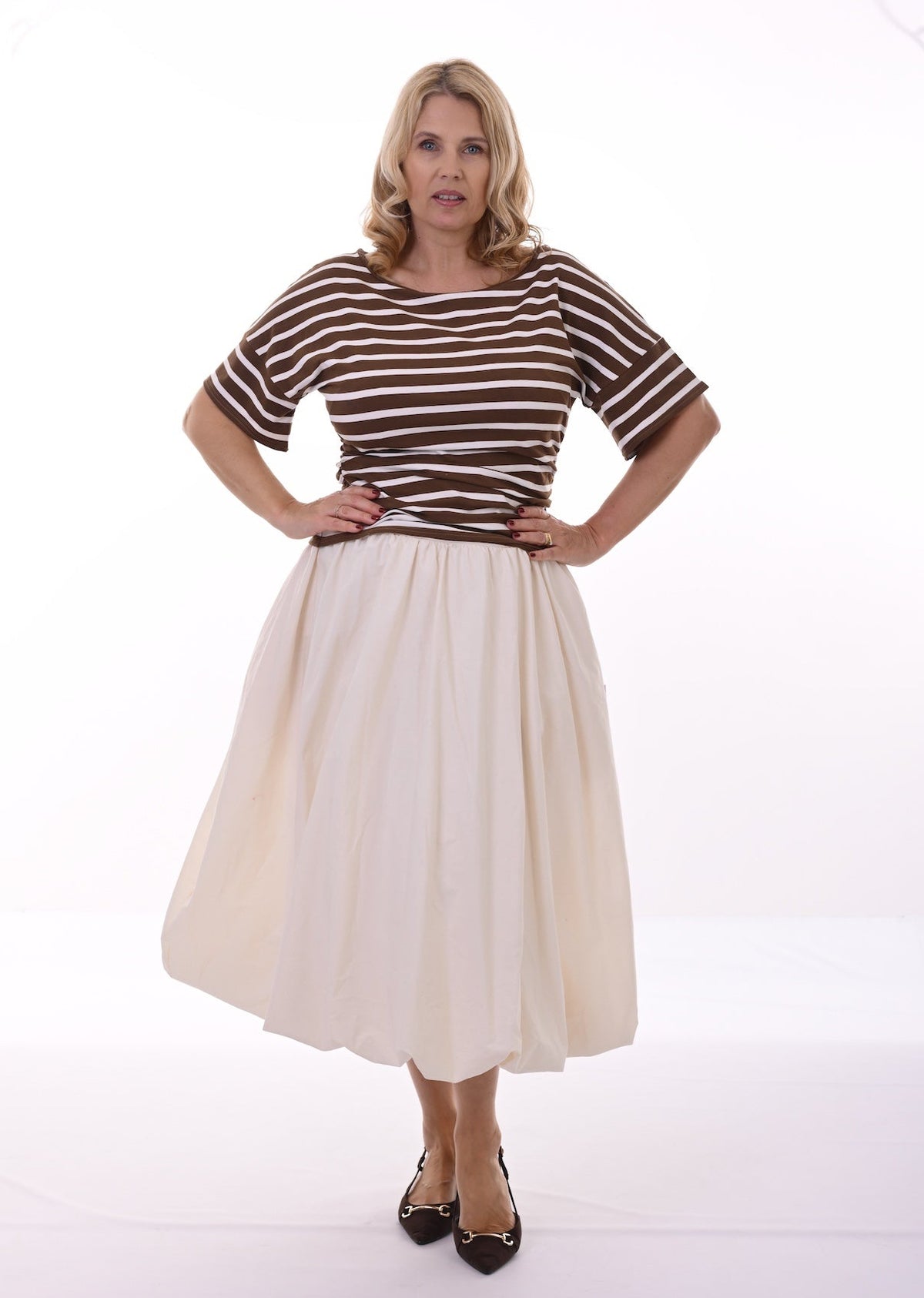 A woman modeling a retro-style outfit consisting of a brown and white horizontally striped short-sleeve top and a pleated cream-colored midi skirt, paired with black pointed-toe flats, against a plain white studio background.