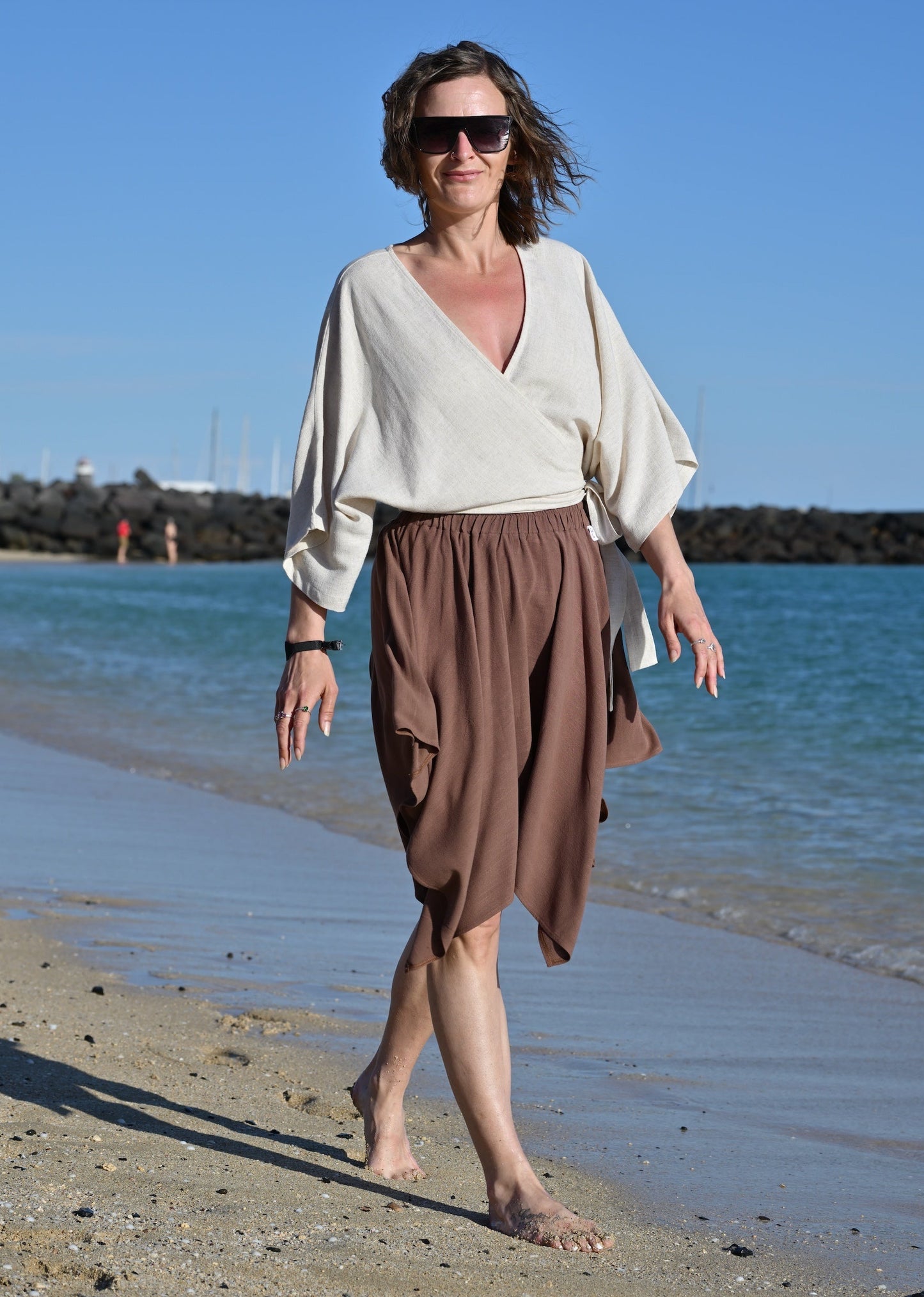 Woman walking on a beach wearing a white top and brown skirt.