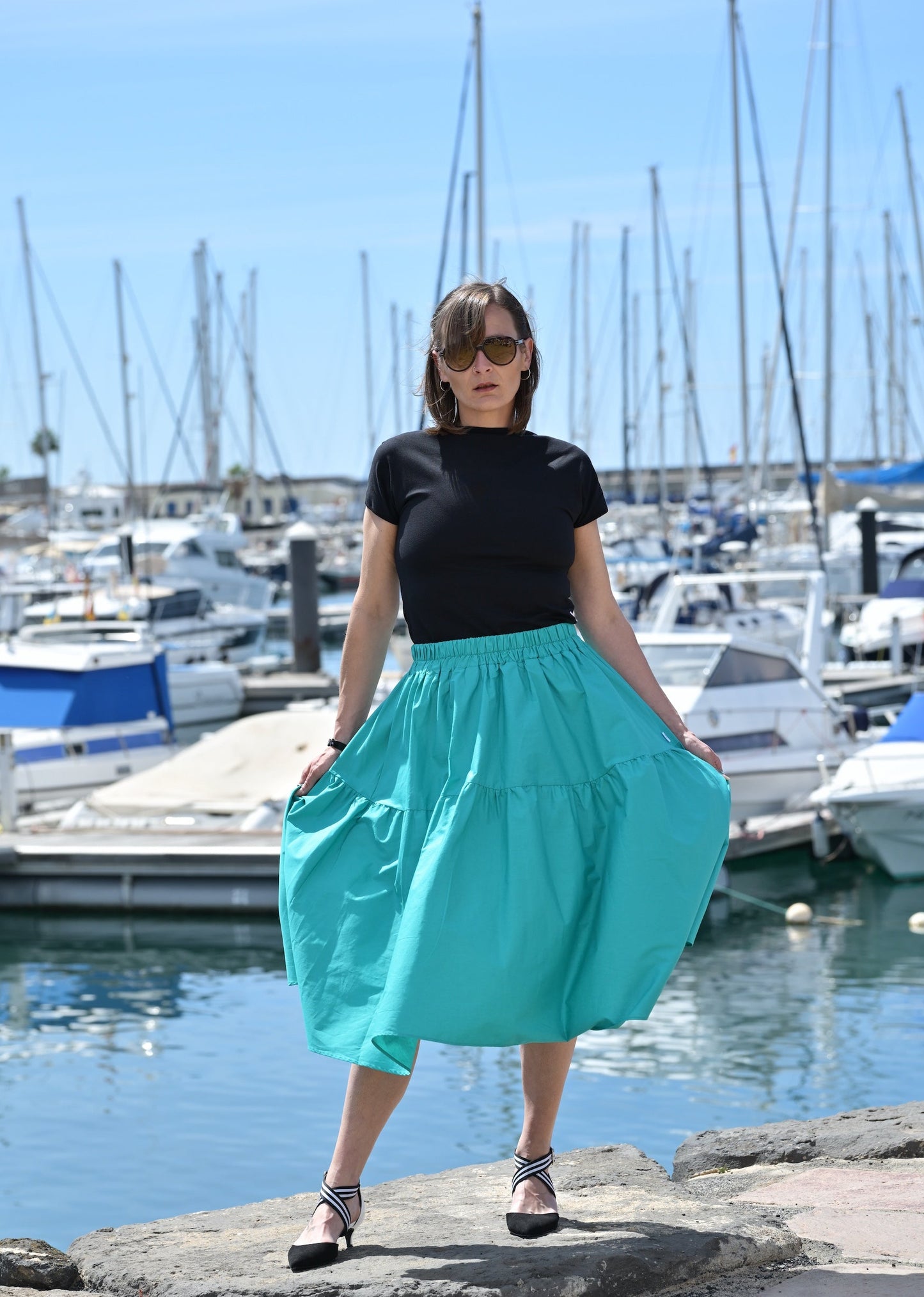 Woman in a black top and teal skirt standing by a marina with boats and clear blue sky.