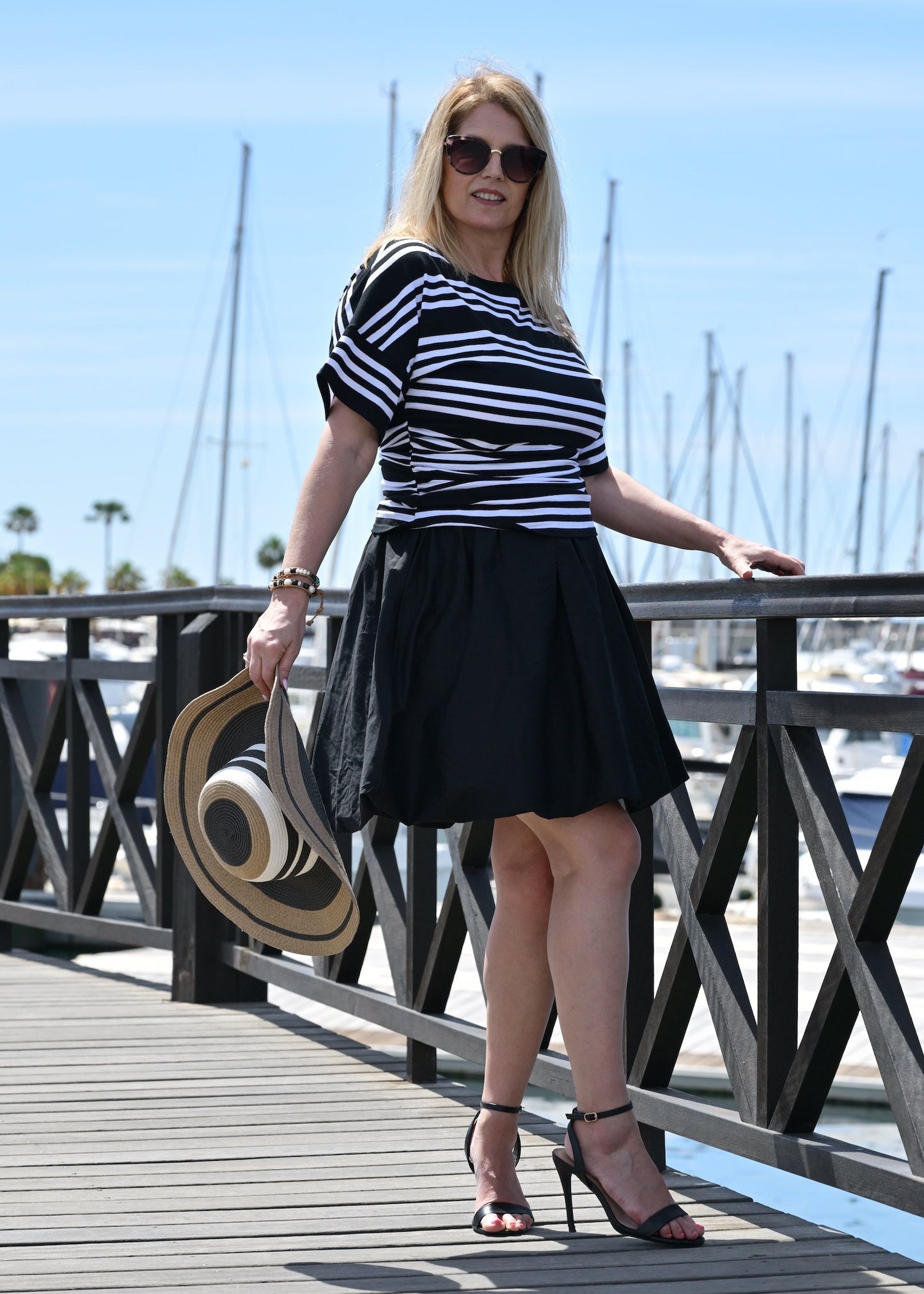 Woman in a striped shirt and black skirt standing on a wooden dock with boats in the background.