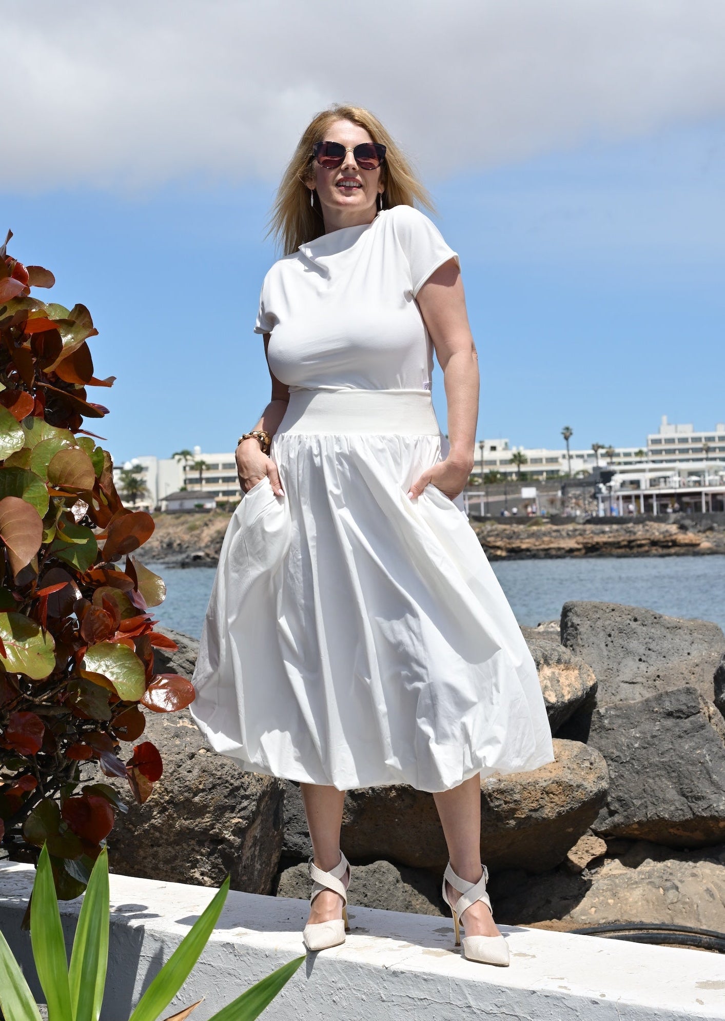Woman in a white dress standing by a waterfront with plants and rocks in the foreground.