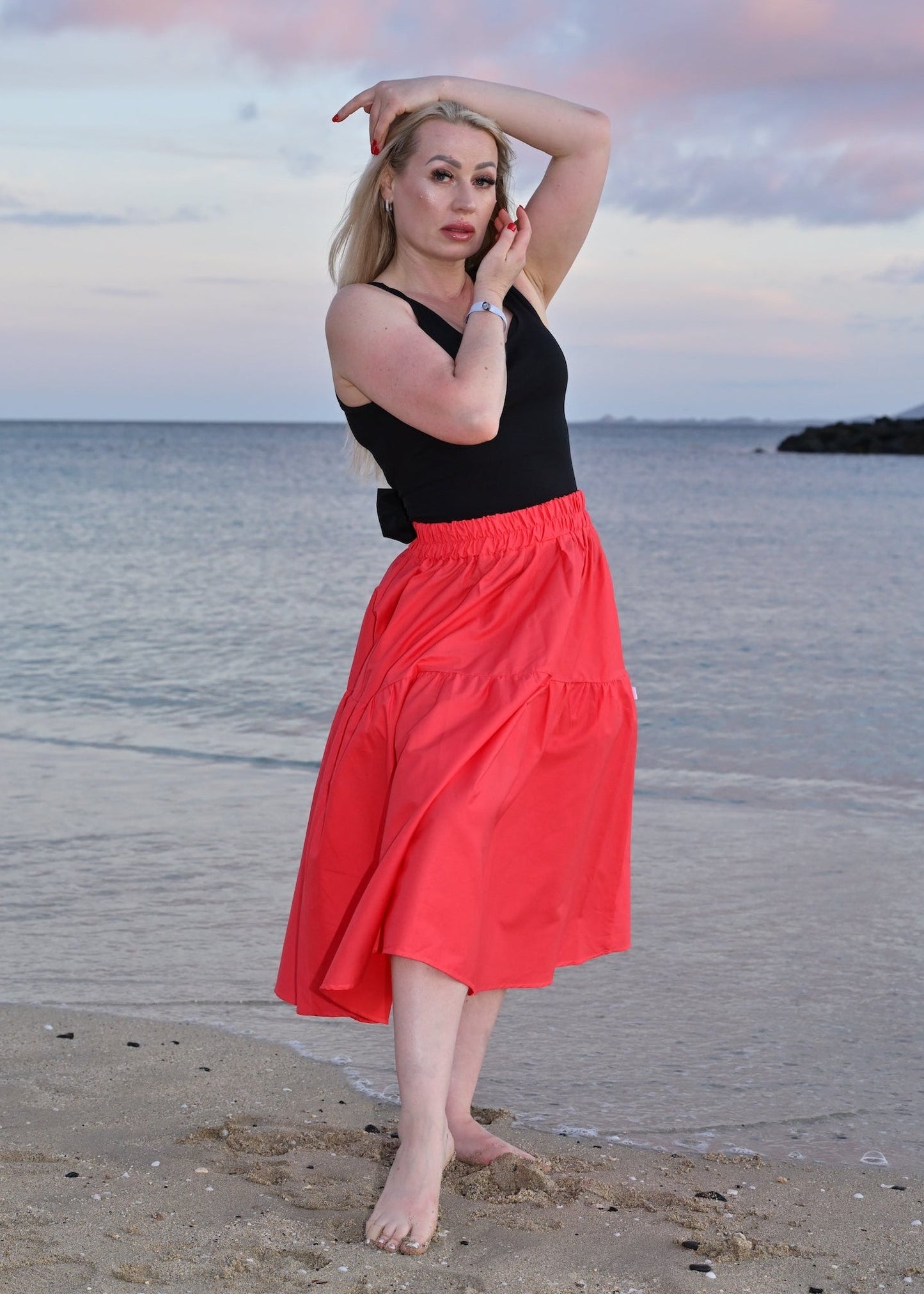 Woman in a black top and coral skirt standing on a beach with water and sky in the background
