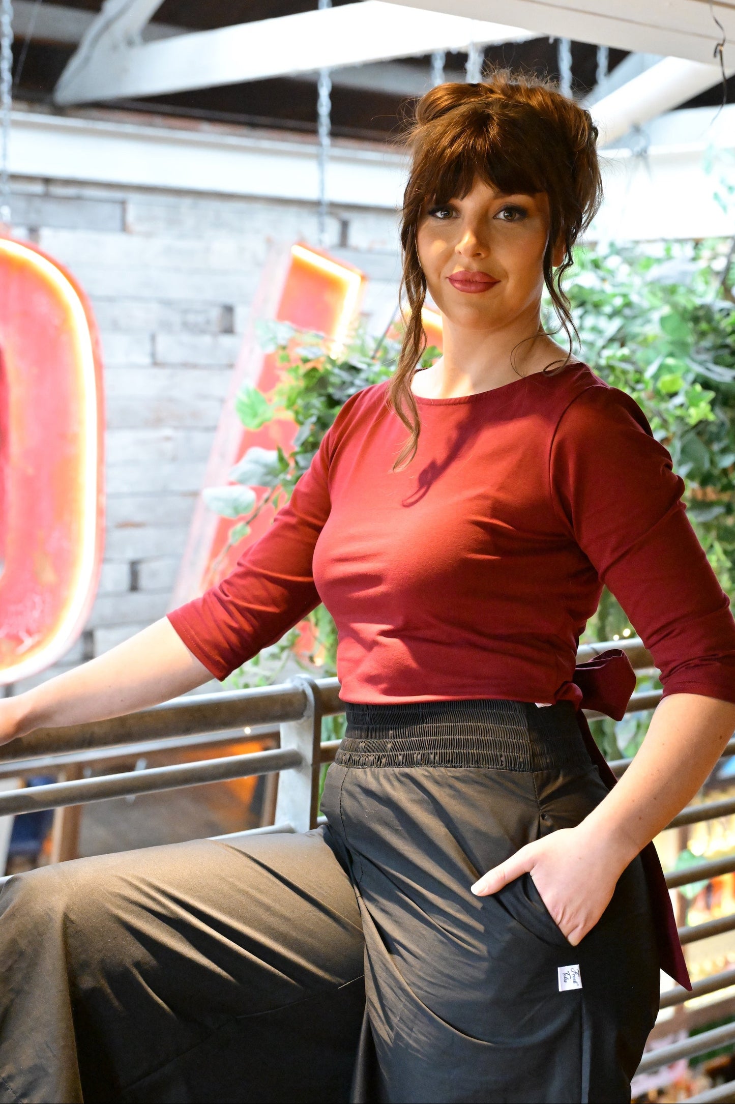 Woman in a red top and black pants sitting on a metal railing with a blurred indoor background