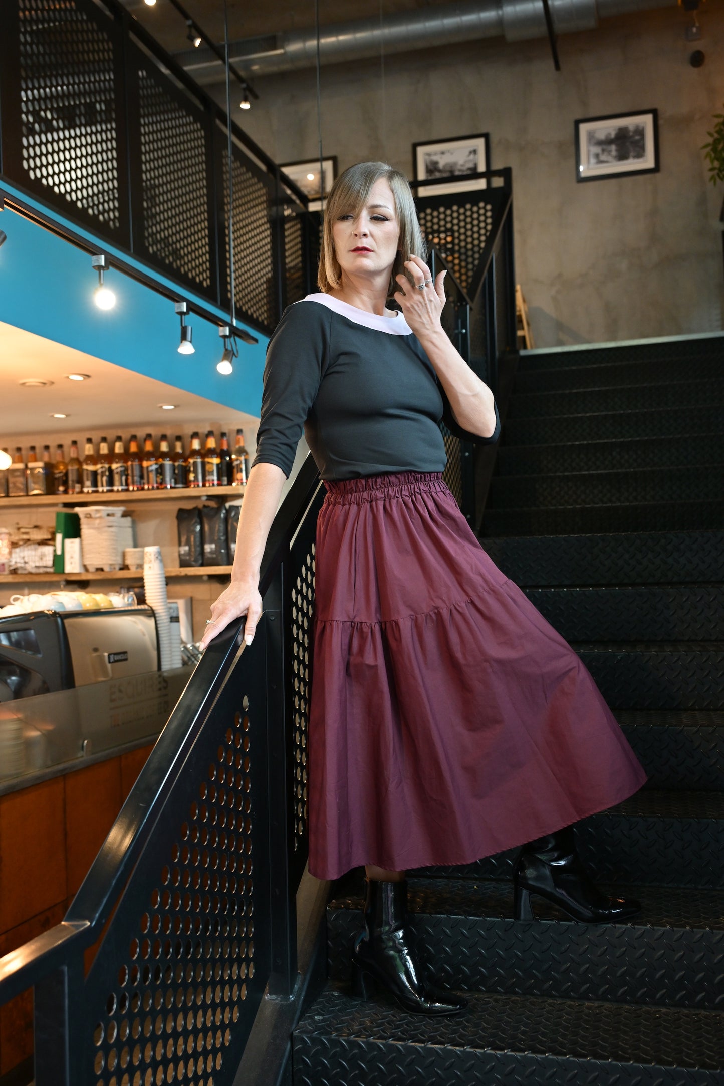 Woman in a maroon skirt and black top standing on a staircase in an indoor setting.