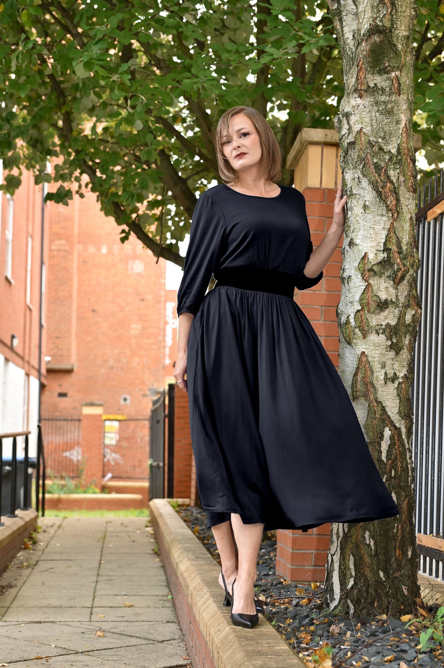 Woman in a black dress standing next to a tree on a sidewalk.