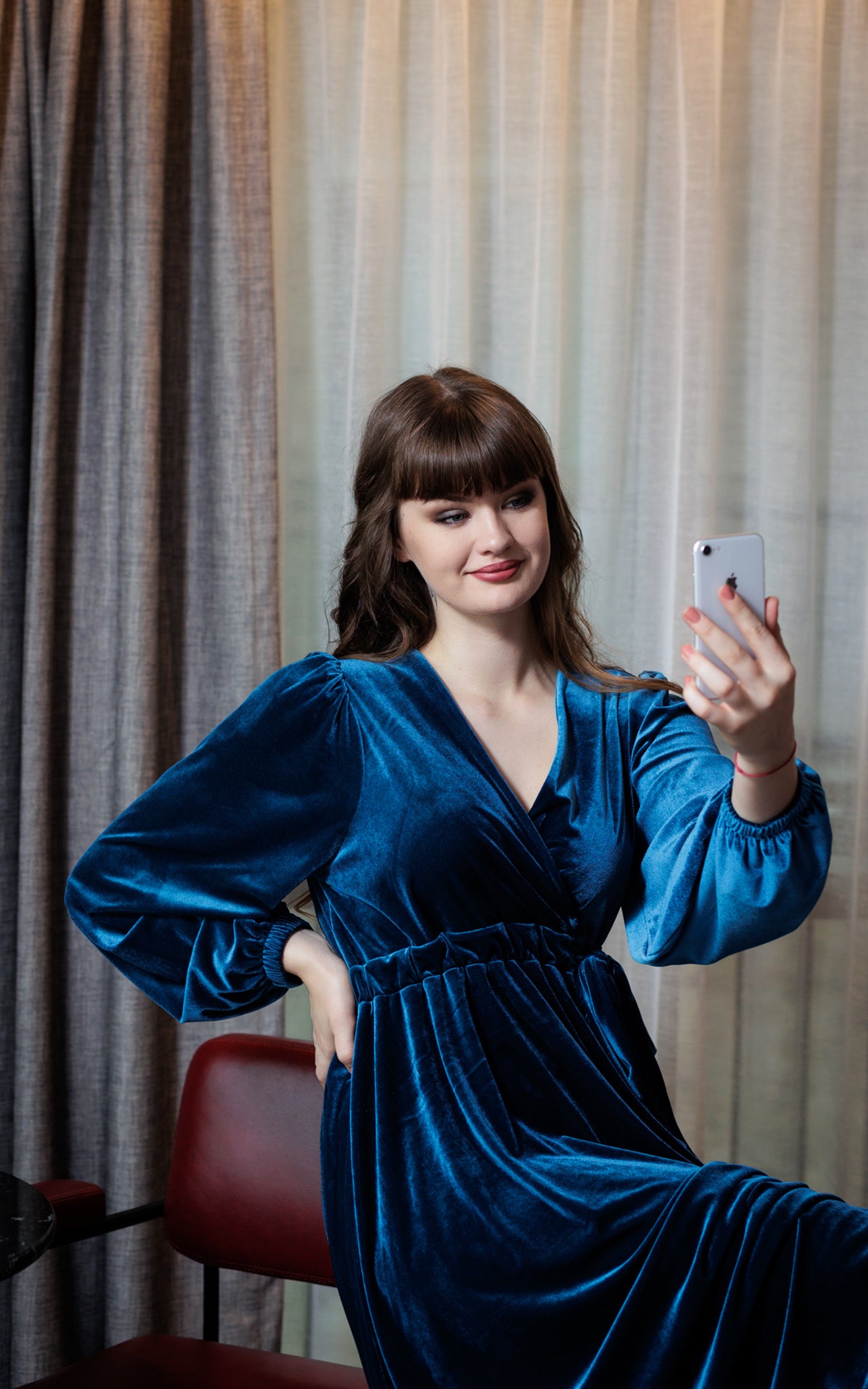 This image is the model seated and taking a photograph with their phone. The model has their hand on their upper waist waist which demonstrated the volume of the sleeve of the teal blue velvet dress she is wearing.. The background is cream and grey. The model has brown hair and is seated on a maroon coloured seat.