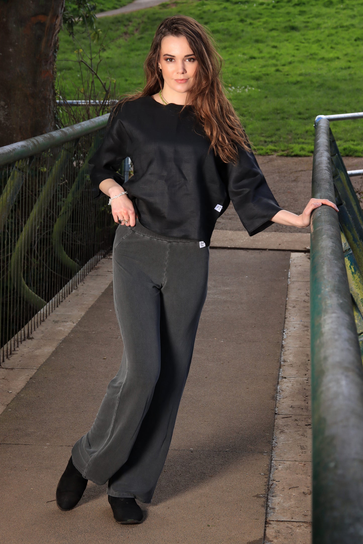 This in a young lady in the centre of the image facing forward with her left hand on a grey metal railing. Her hair is long and brown and it falls mainly over her left shoulder. She is wearing a black top with 3/4 sleeves and round neck. Her trousers are charcoal grey.