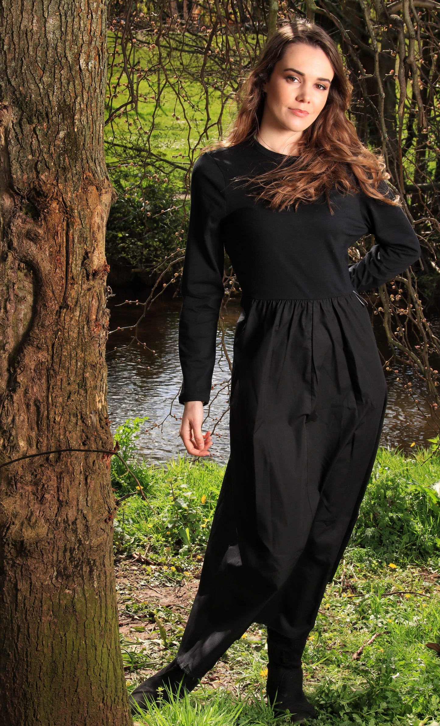 This is a young lady wearing a midi/maxi length black dress with a round neck, long sleeves and a gathered skirt. She is wearing black boots and silhouetted against a countryside background, next to a stream.