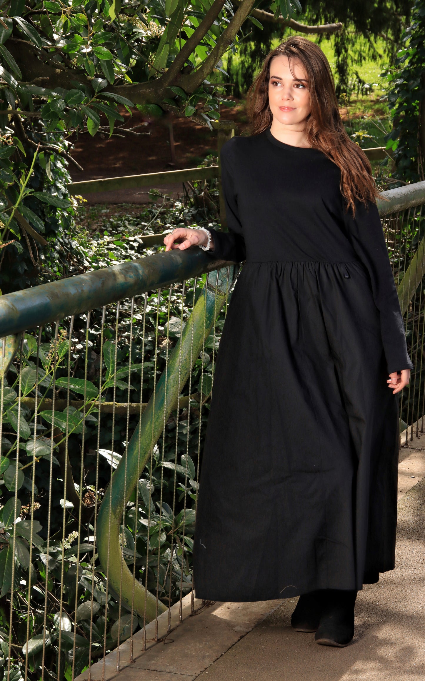 This is a young lady wearing a midi/maxi length black dress with a round neck, long sleeves and a gathered skirt. She is wearing black boots and silhouetted against a woodland background, leaning against a bridge.