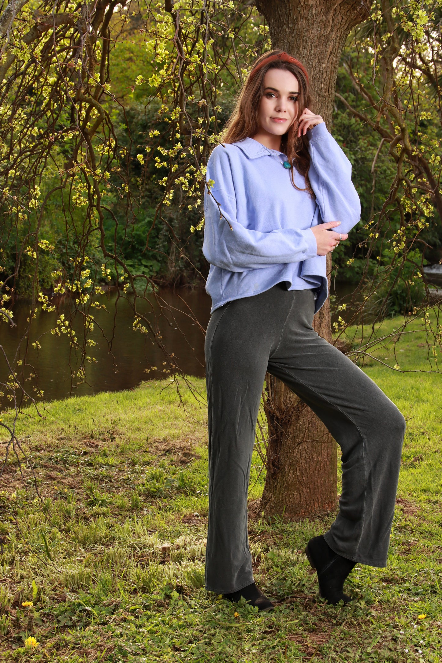 This is an image of a young lady standing in front of the truck section of a tree. She faces to the right of the image and is supporting the left elbow with her right hand. Her left leg is bent. She wears a lilac sweatshirt with a collar and charcoal grey pants.