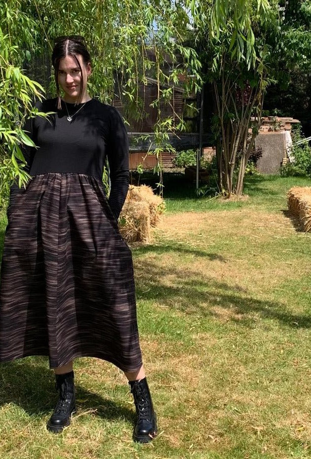 This is a young woman standing in a country garden. She has dark hair which is tied up and is facing the camera with her hands in her pockets. She is wearing black boots. She is wearing a midi dress with a black bodice and a full black and tan printed skirt.