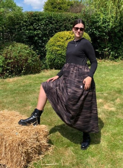 This is a young woman standing in a country garden. She has dark hair which is tied up and is facing the camera with her right foot resting on a straw bale. She is wearing sunglasses with black boots. She is wearing a midi dress with a black bodice and a full black and tan printed skirt.