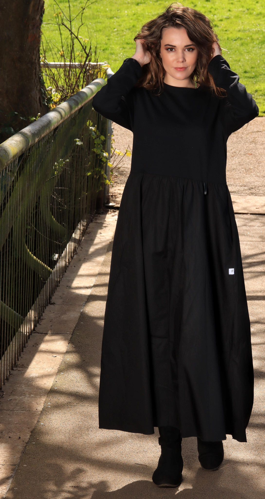 This is a young woman with shoulder length dark hair wearing a midaxi black dress with long sleeves. She is silhouetted against a countryside background and is facing the camera. She is wearing black boots.