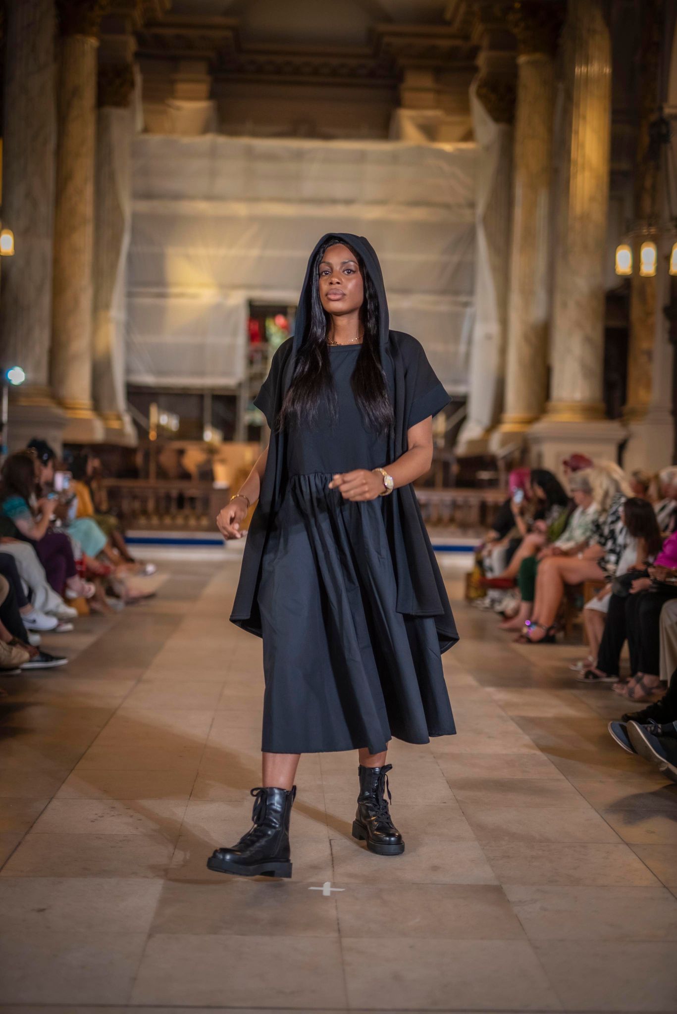 The model is standing in the centre of the pic wearing a black dress. This is a fashion show and the people are sitting in a line at either side of the stone catwalk which is on the ground. The model is holding the hood of the longline Moonrise Shrug which is also black. She wears black lace up boots.