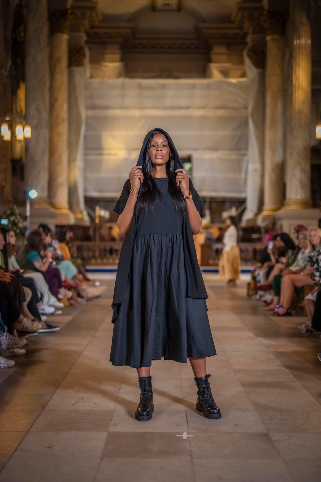 The model is standing in the centre of the pic wearing a black dress. This is a fashion show and the people are sitting in a line at either side of the stone catwalk which is on the ground. The model is holding the hood of the longline Moonrise Shrug which is also black. She wears black lace up boots.