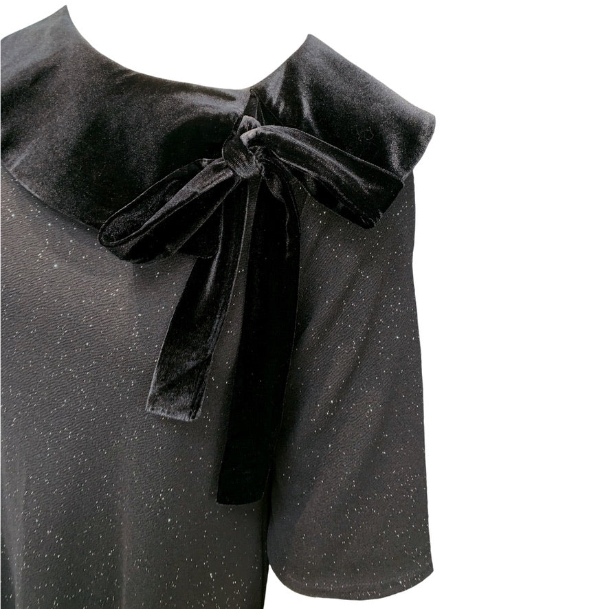 This is a close up of a dress with flared sleeves. The main body of the dress is black with silver sparkles, and a contrasting black velour collar lays across the shoulders, tying into a bow on one side.