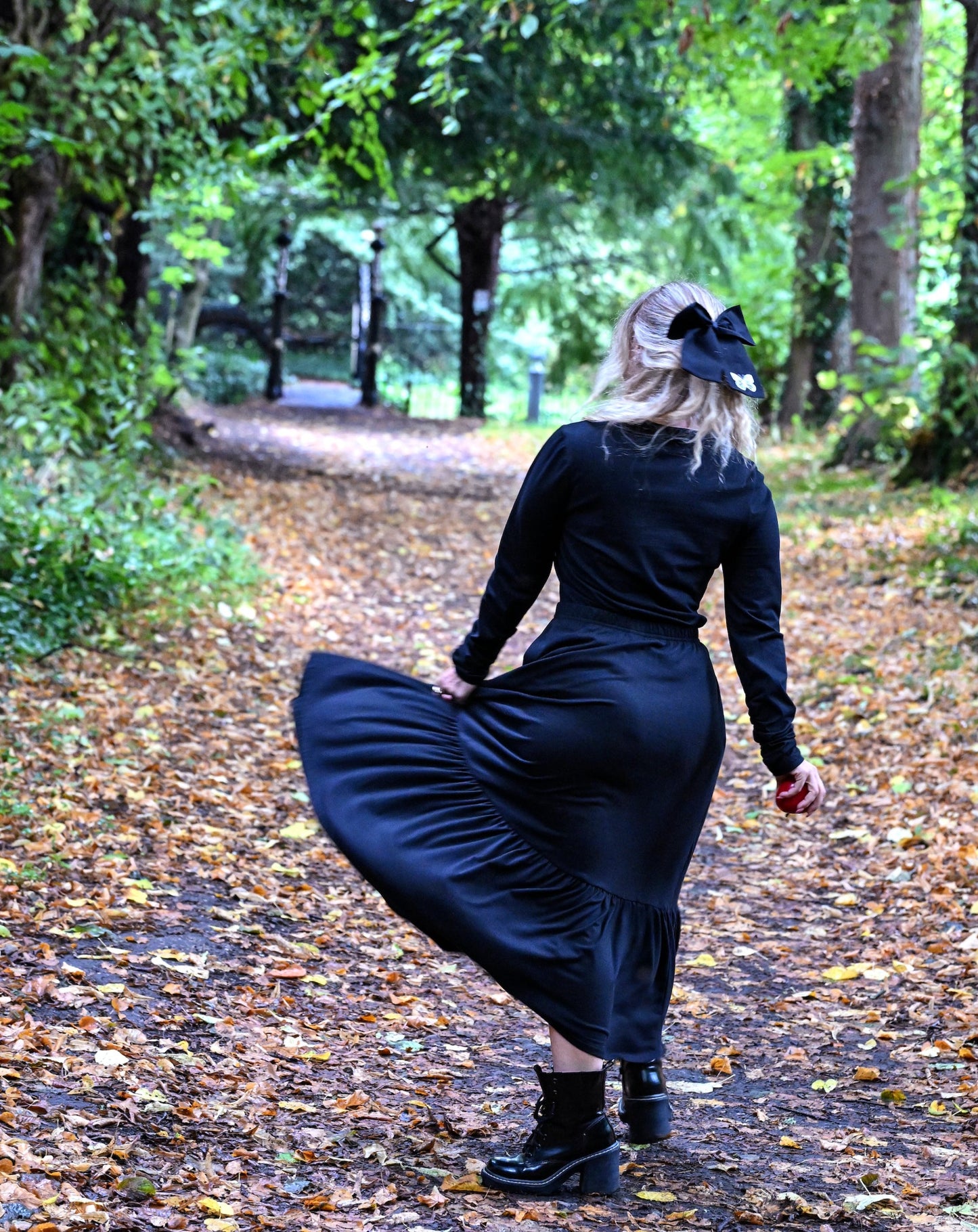 The young woman in the picture has long blonde hair with a black bow. She is walking along a leaf covered path in a park with lots of trees and foliage. She is holding her skirt with her left hand. She has her back to the camera and she is holding an apple in her right hand. She is wearing a beatnik top with crew neck in black. The top has ruched sleeves and is being worn with our boho black skirt and black boots.