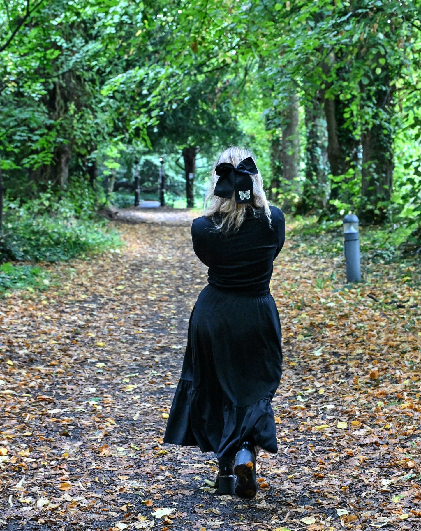 The young woman in the picture has long blonde hair tied with a black bow. The bow has a white frock tales butterfly on it. She is walking along a leaf covered path in a park with lots of trees and foliage. She has her back to the camera and she is holding her arms in front of her. She is wearing a beatnik top with crew neck in black. The top has ruched sleeves and is being worn with our boho black skirt and black boots.