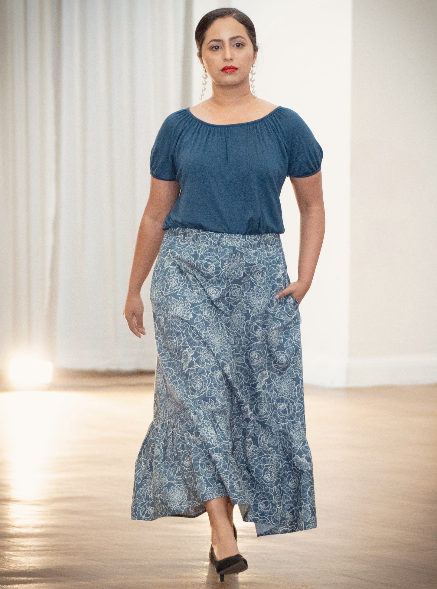 The young woman in the picture has dark hair and she is walking towards the camera in a well lit room. She is wearing a boho gathered midi skirt with pockets in navy print. She is also wearing our posey top in french navy. She is wearing black shoes. Her right hand is by her side while her left hand is in a pocket.