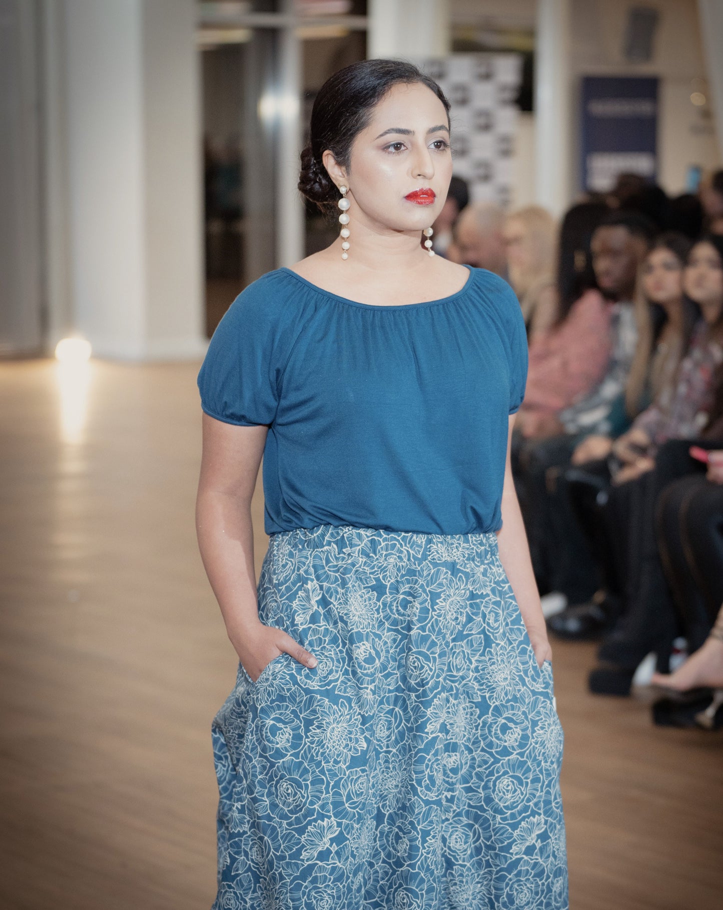 The young woman in the picture has dark hair and she is walking forward in a crowded room. She has long silver earrings and red lipstick. She is wearing a boho gathered midi skirt with pockets in navy print. She is also wearing our posey top in french navy. Both hands are in her pockets.  