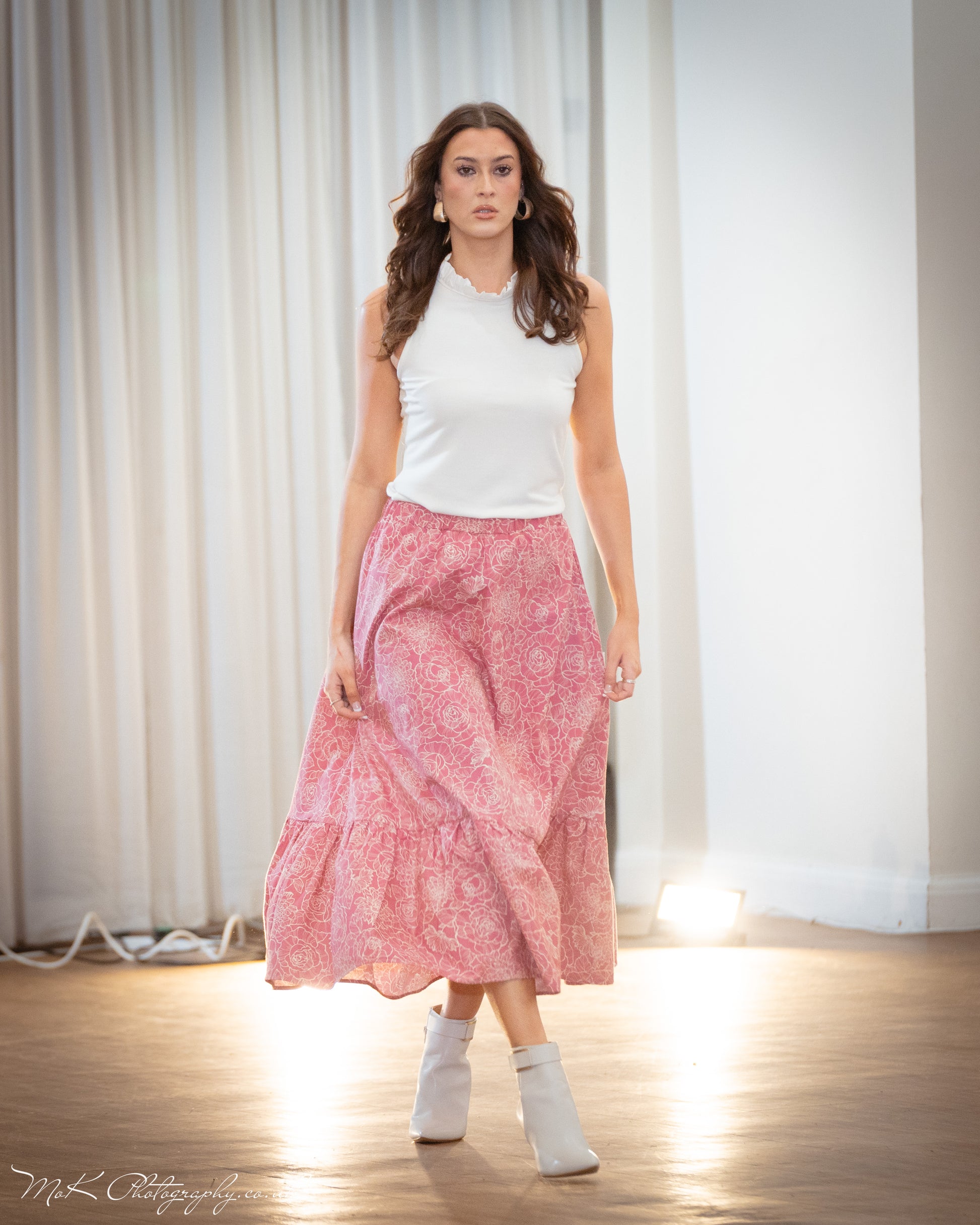 This is a young woman walking towards the camera. She is wearing a pink and cream patterned skirt silhouetted against a white drape background. It has an elasticated waist and frill hem. She wears this with a cream sleeveless top with ruffle neck.