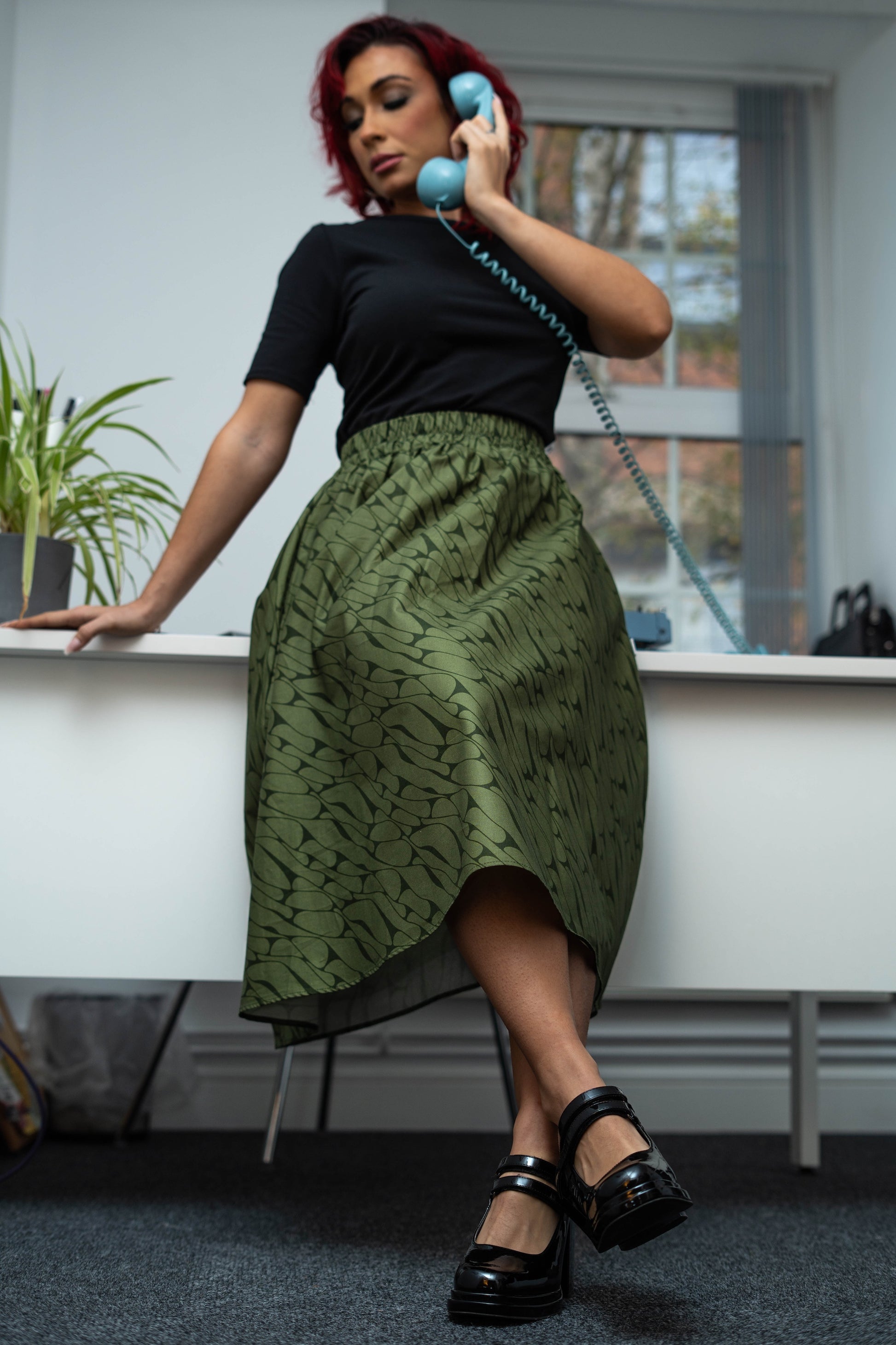 Person wearing a green patterned skirt and black top, holding a blue phone receiver indoors.